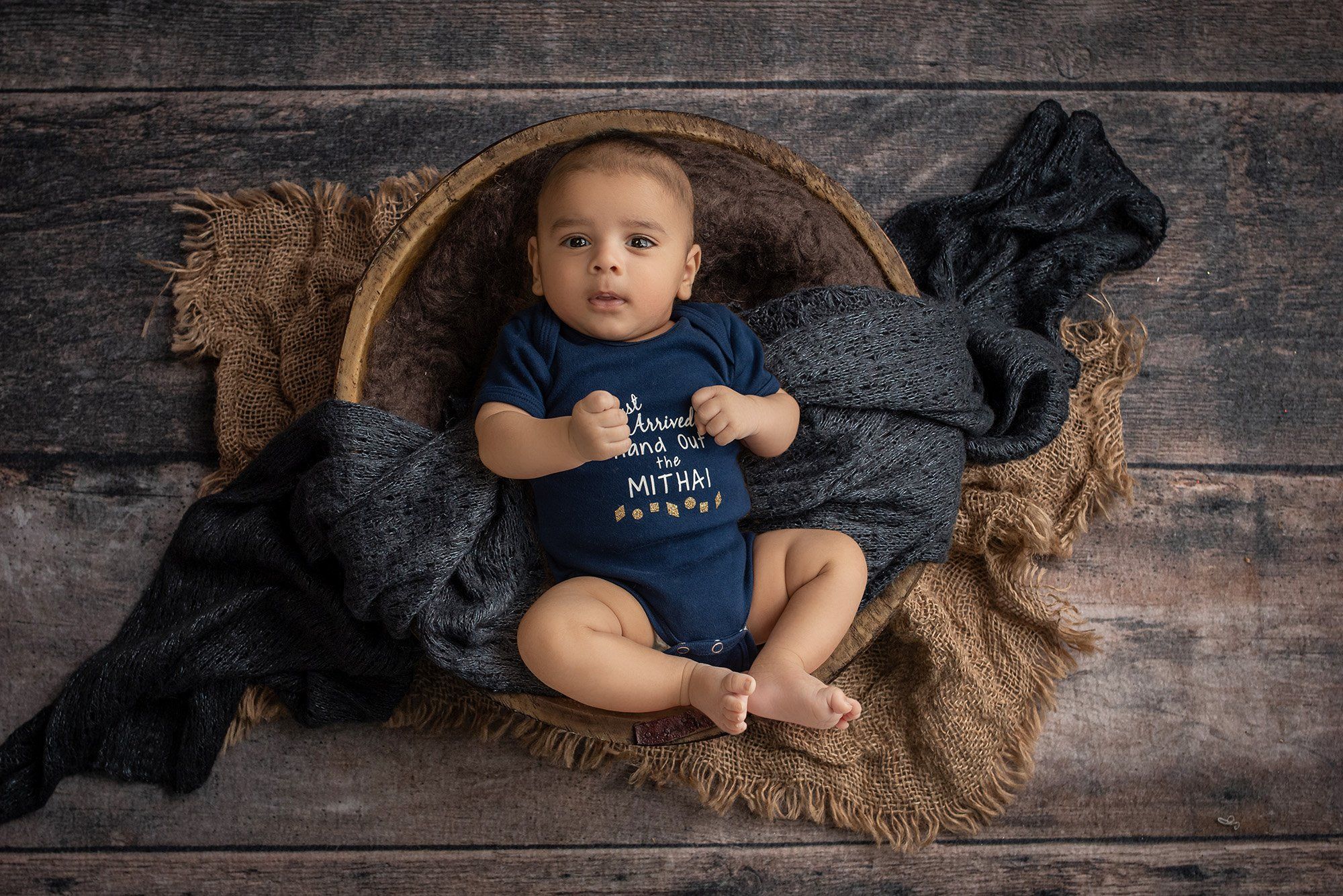 newborn baby boy wearing navy blue onesie laying inside wooden bowl with blue and brown blankets