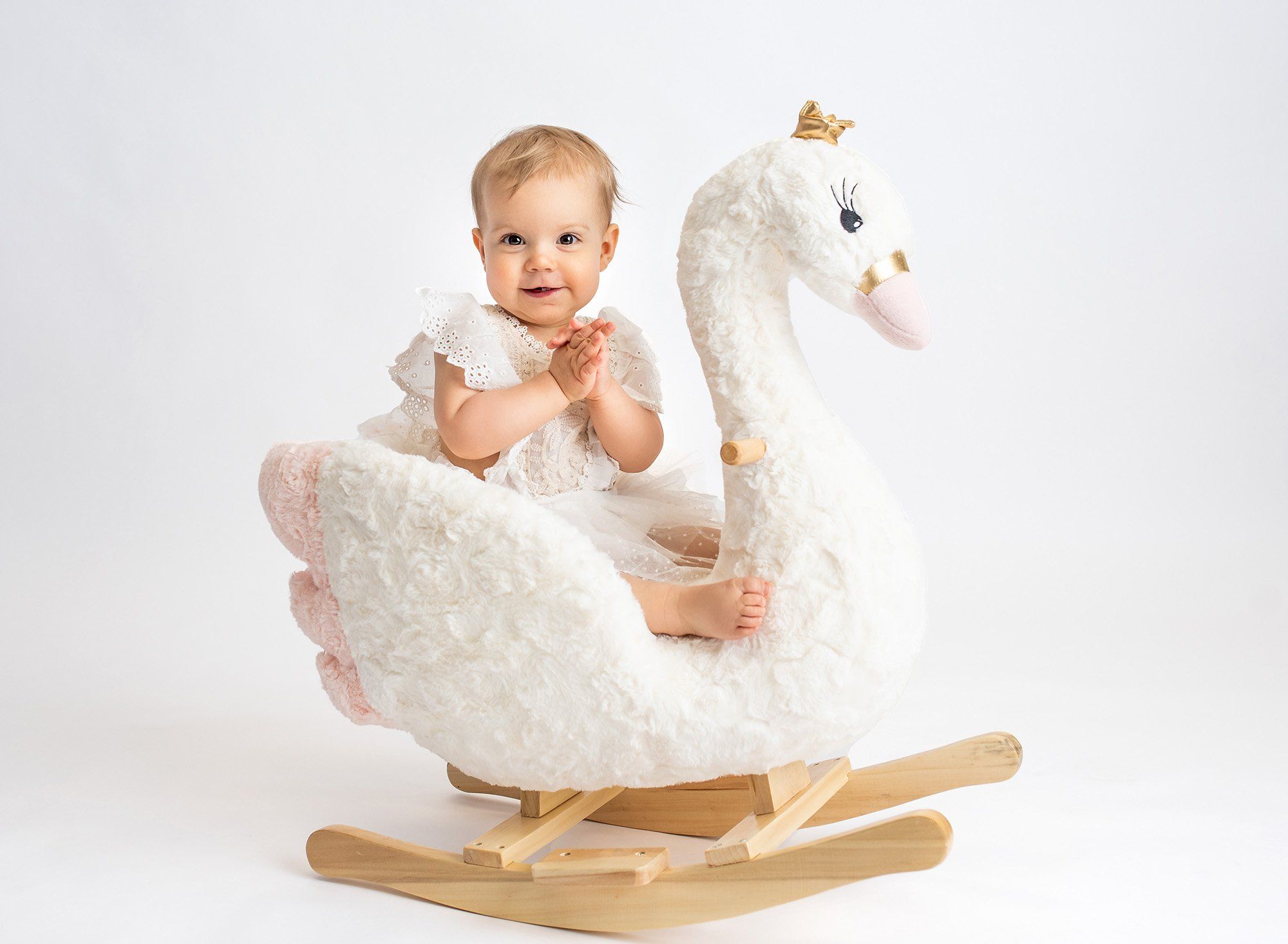 one year old girl wearing white lace dress sitting on swan rocker with white backdrop