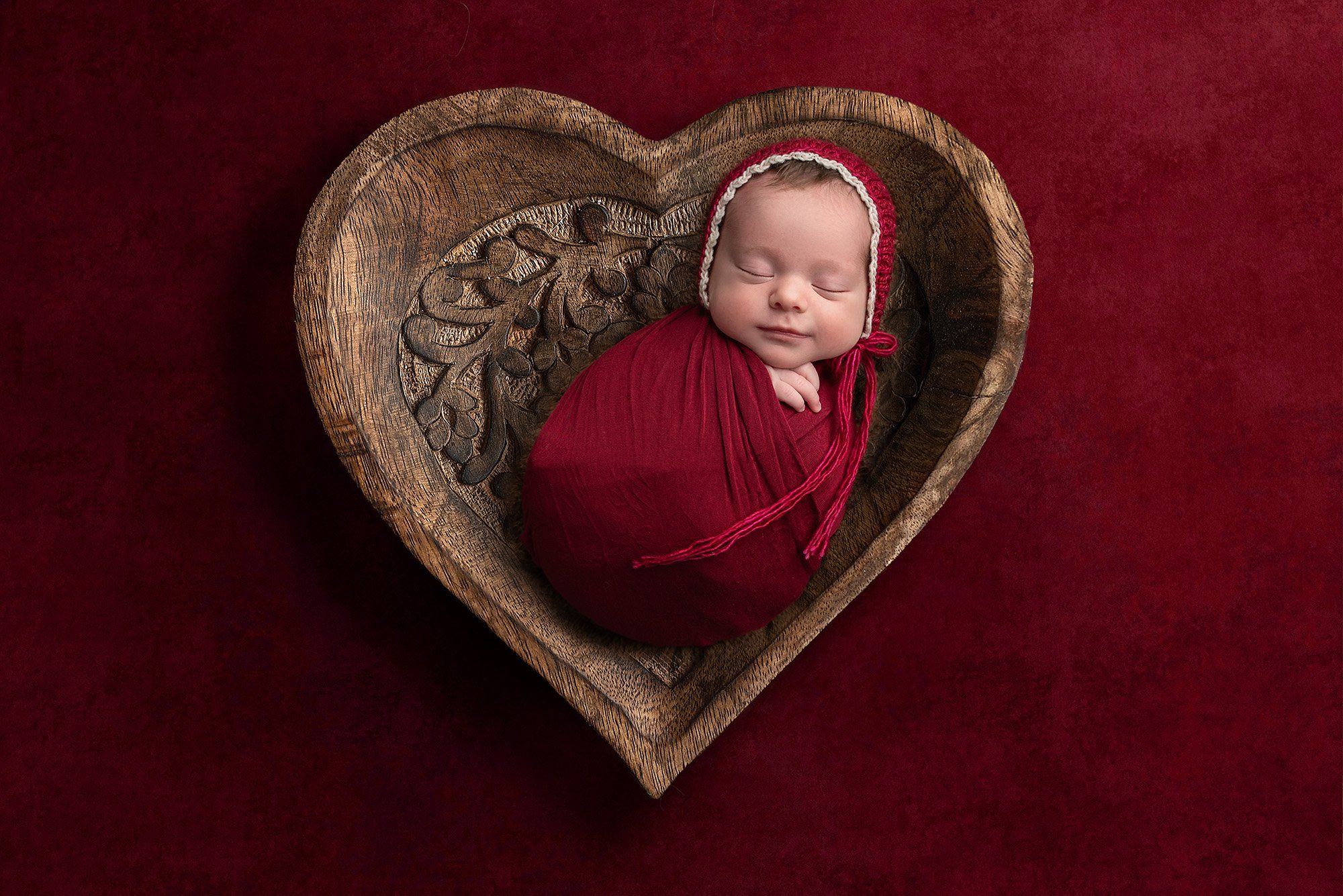 newborn baby girl sleeping wearing red bonnet swaddle laying in wooden heart box on red background