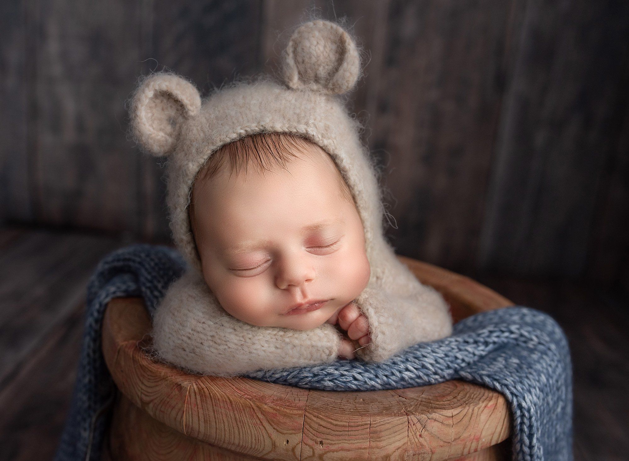 newborn baby boy dressed in bear sweater romper sitting in wooden bowl with blue sweater blanket