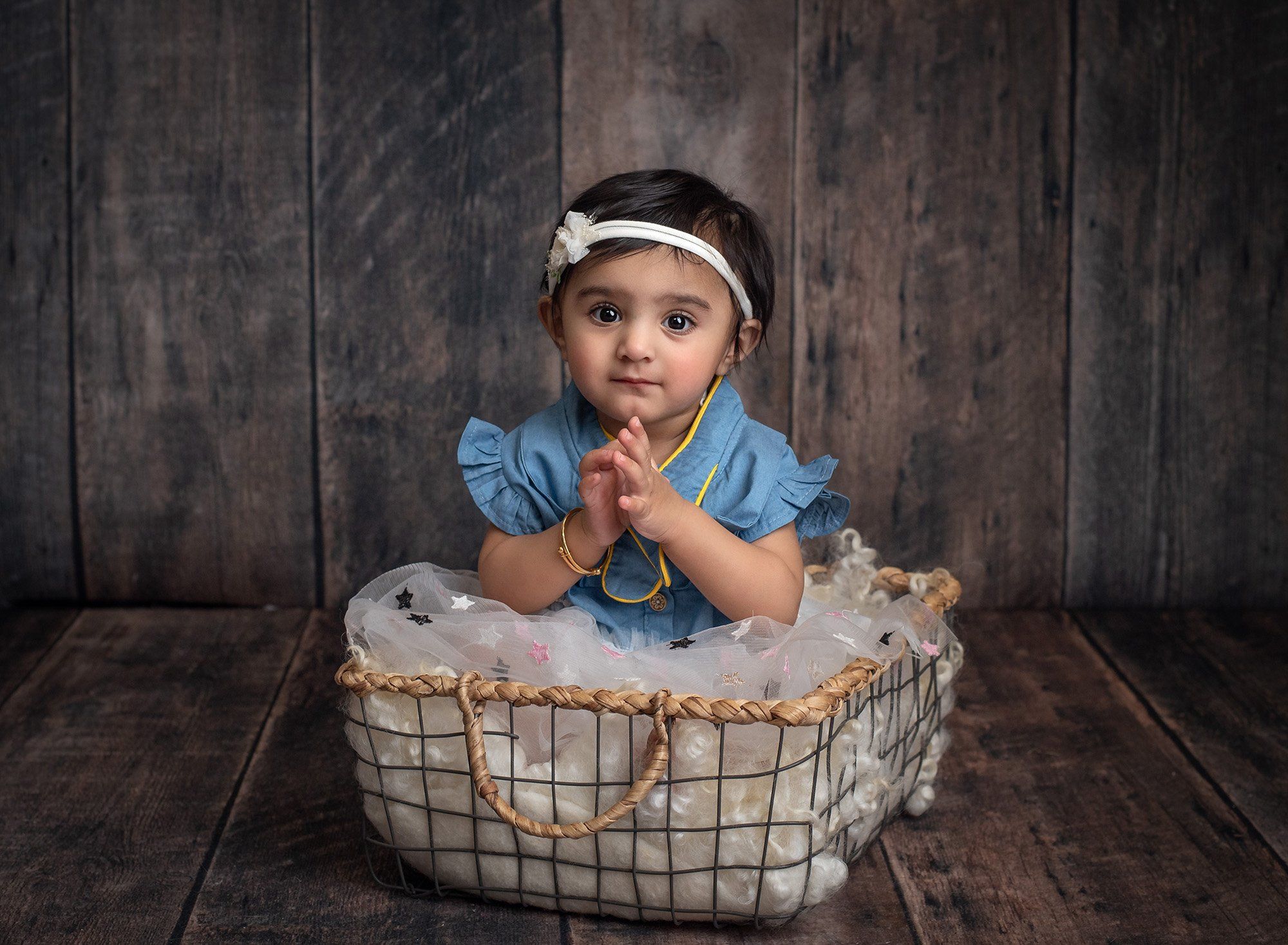 baby girl sitting in black wire basket with ruffled star blanket wearing blue dress and white headband on wooden backdrop