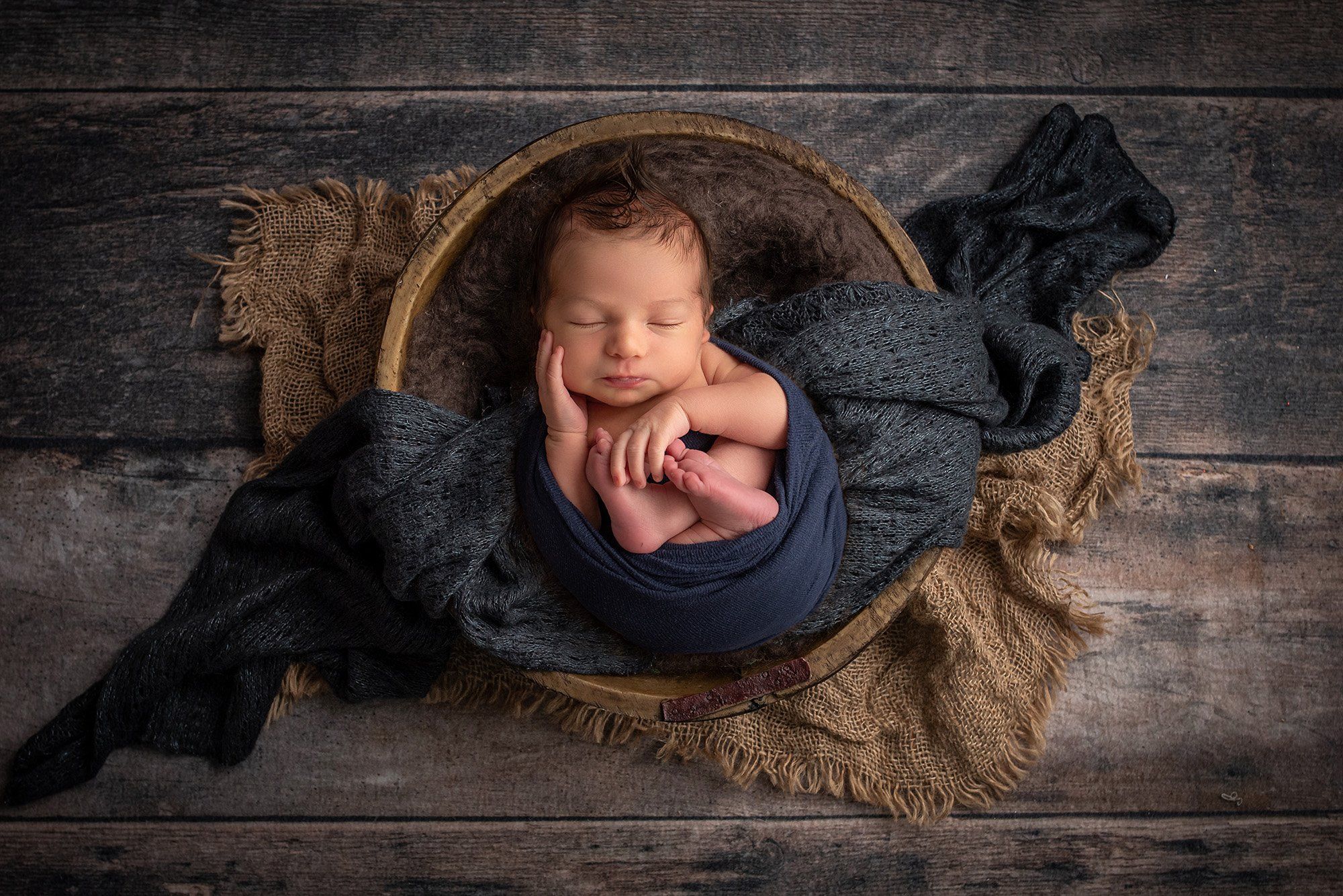 newborn baby sleeping in wood bowl wrapped in navy blue cotton wrap with wooden background
