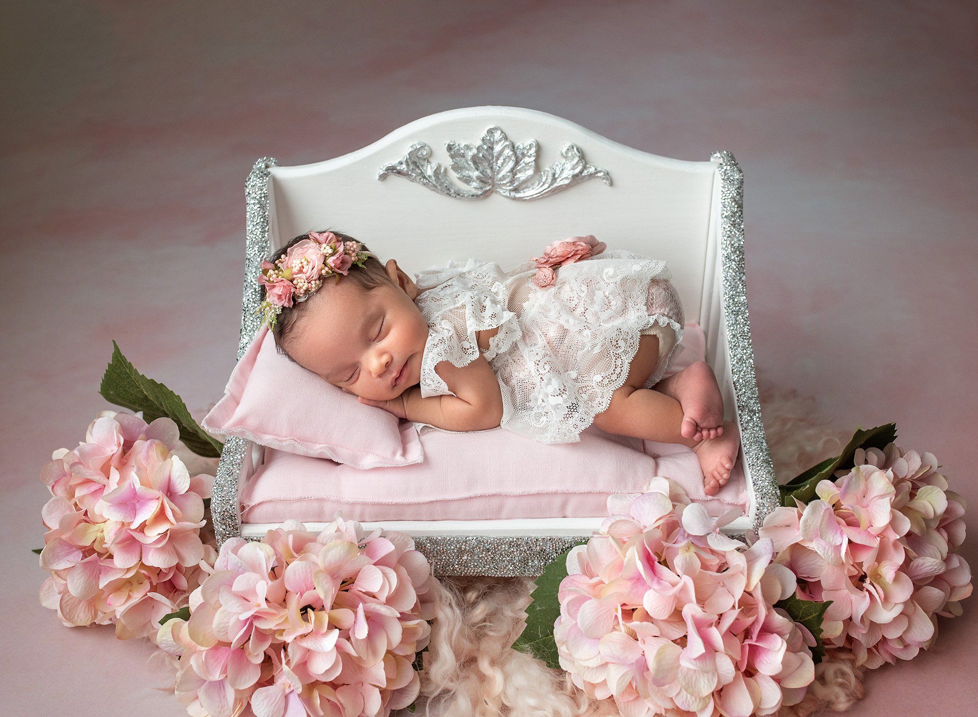 newborn girl wearing lace sleeping on miniature bed surrounded by pink flowers