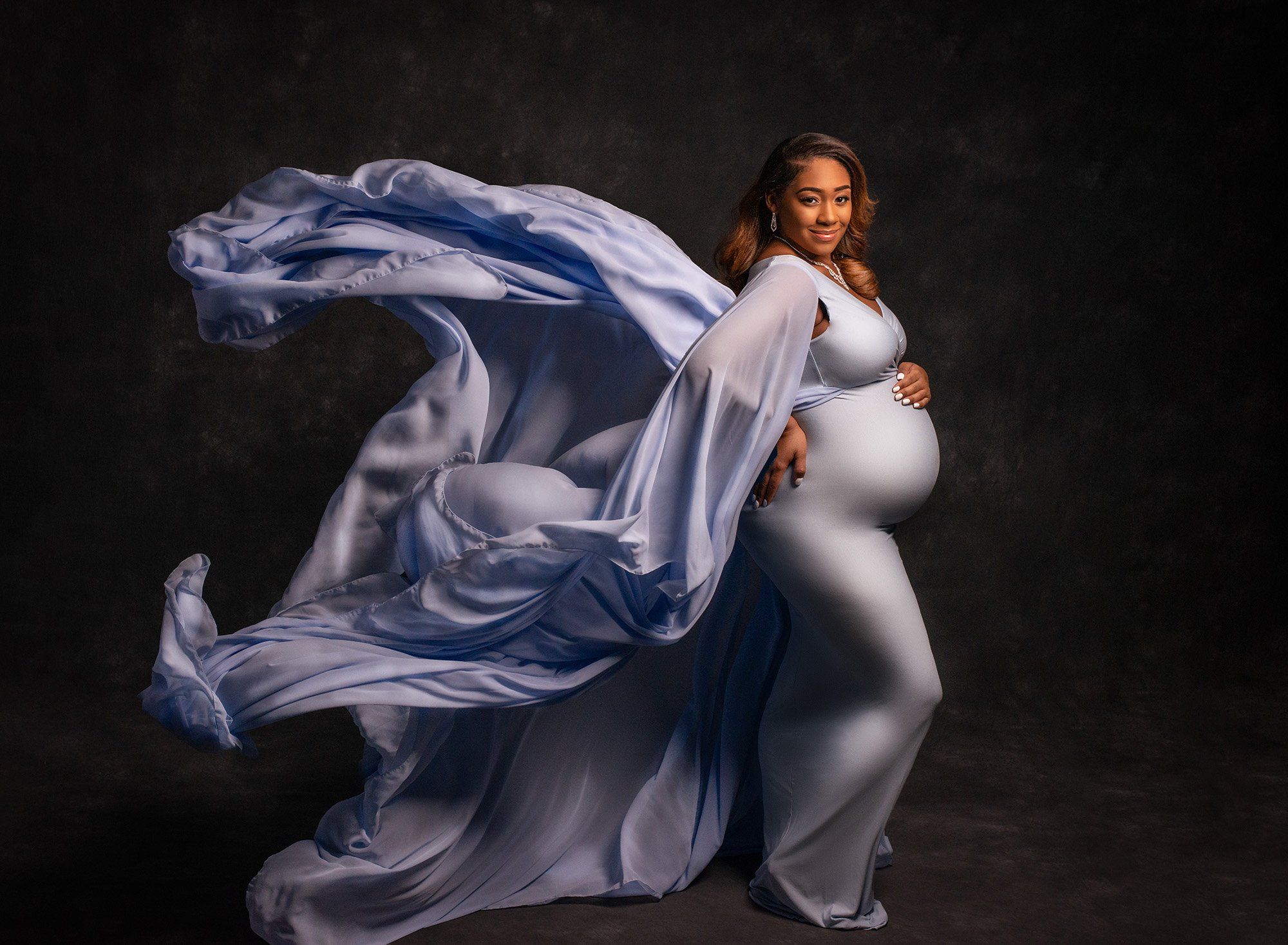 pregnant woman wearing periwinkle flowing maternity gown on dark gray backdrop