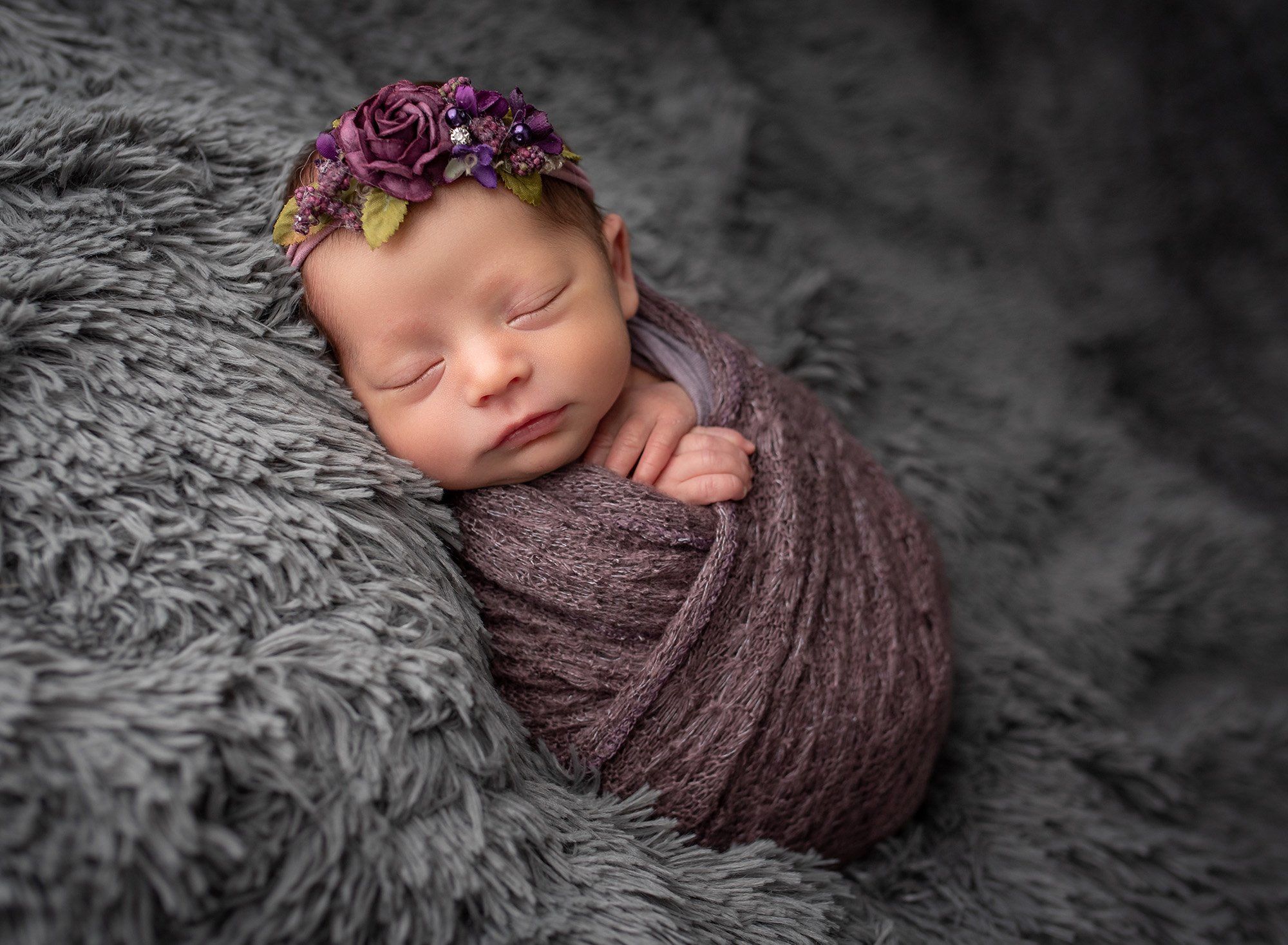 sleeping newborn baby girl on fluffy gray blanket swaddled in purple wearing purple floral headband