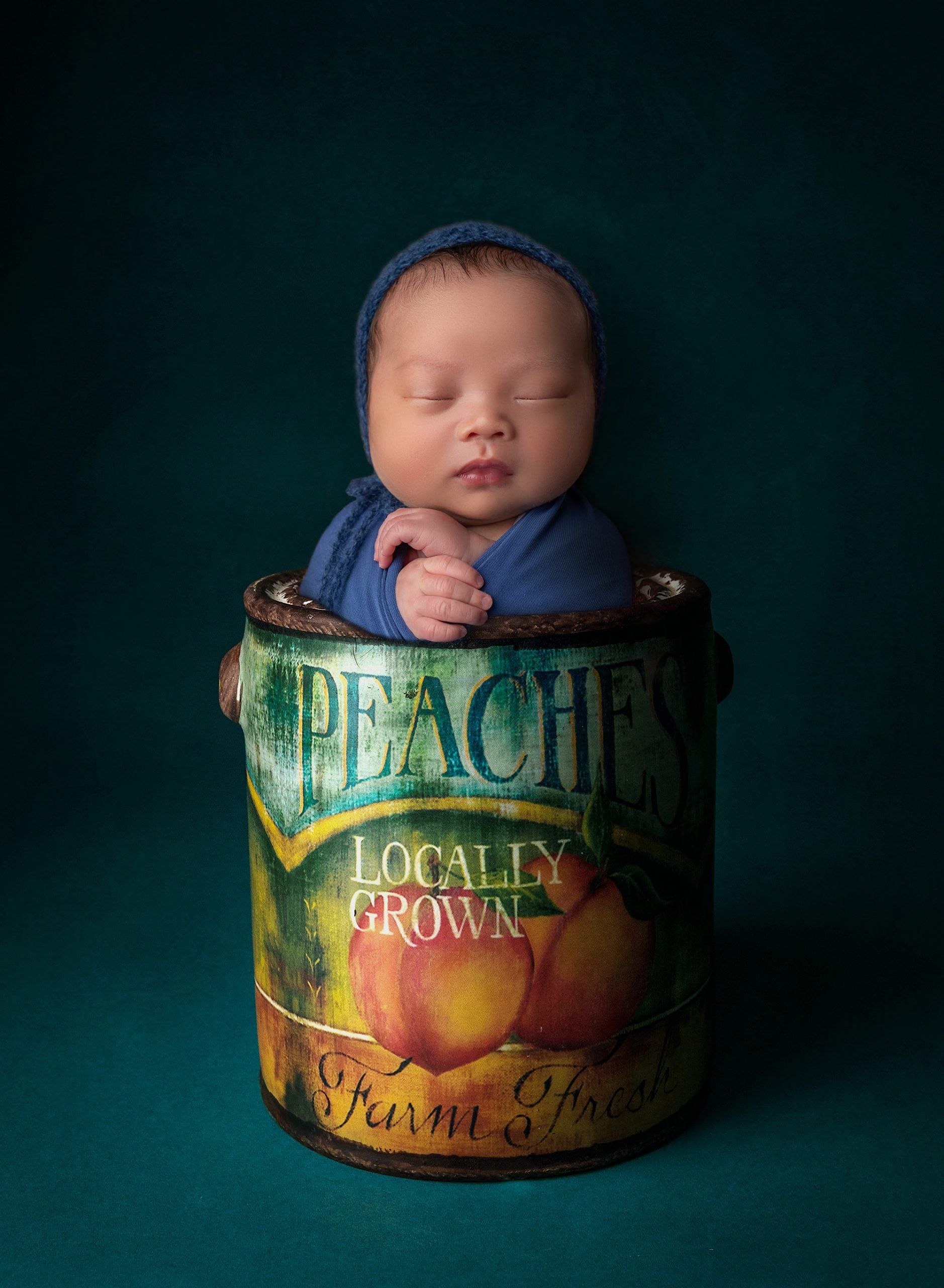 sleeping newborn baby boy swaddled in royal blue wrap sitting out of a peaches farm can