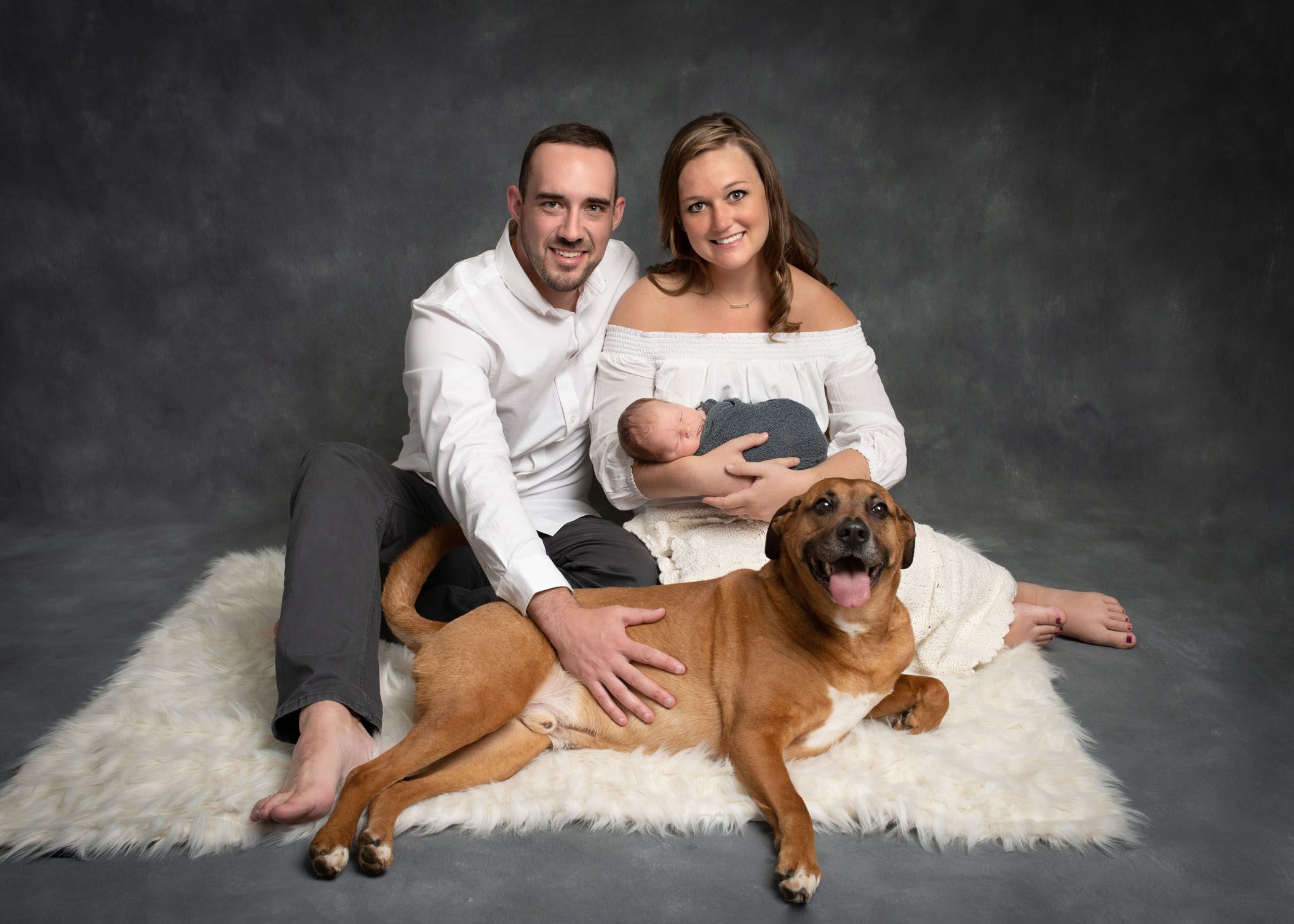 Parents holding a newborn baby with their dog on the floor in front of them