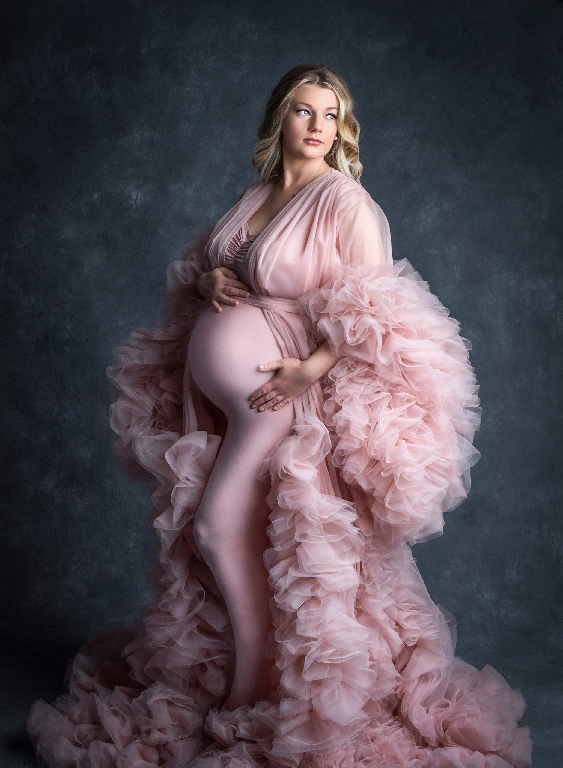 blonde pregnant woman posing in pink ruffled maternity dress on grey background