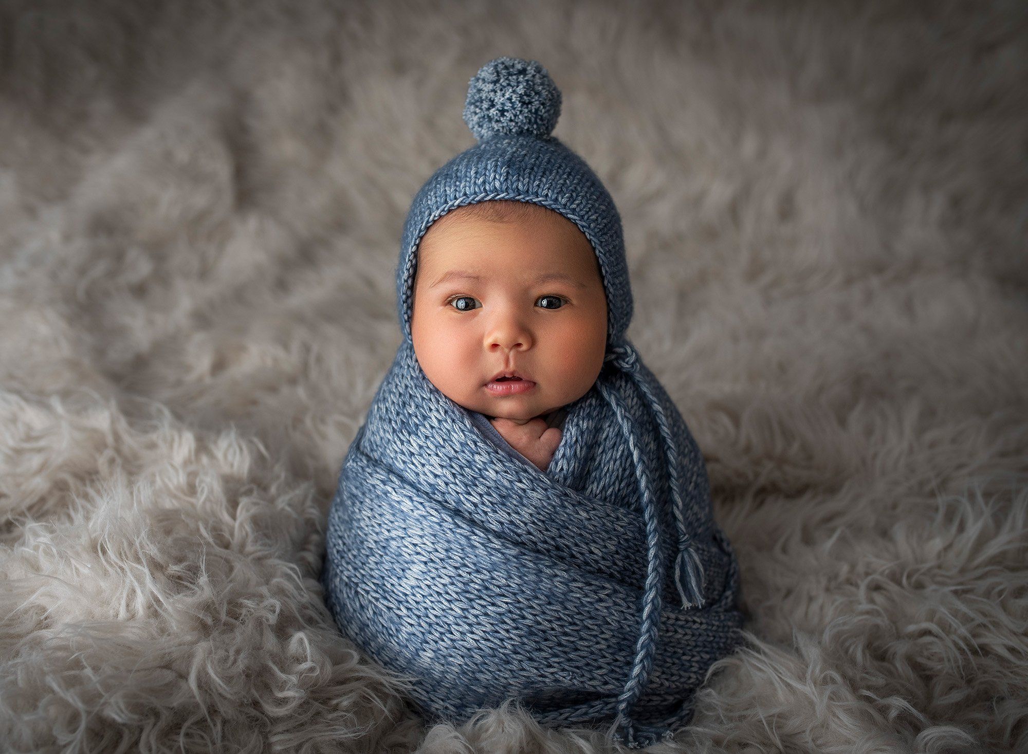 newborn baby boy wide awake swaddled in blue on a cream furry blanket