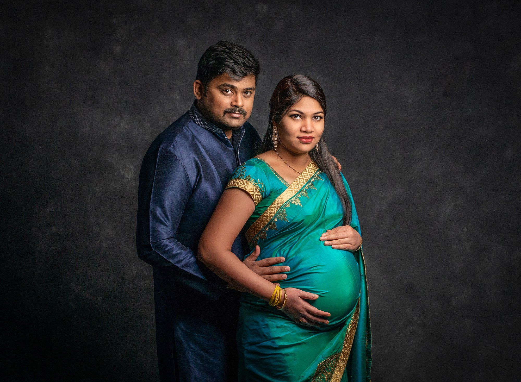 pregnant woman posing with husband in Indian dress clothing on gray backdrop