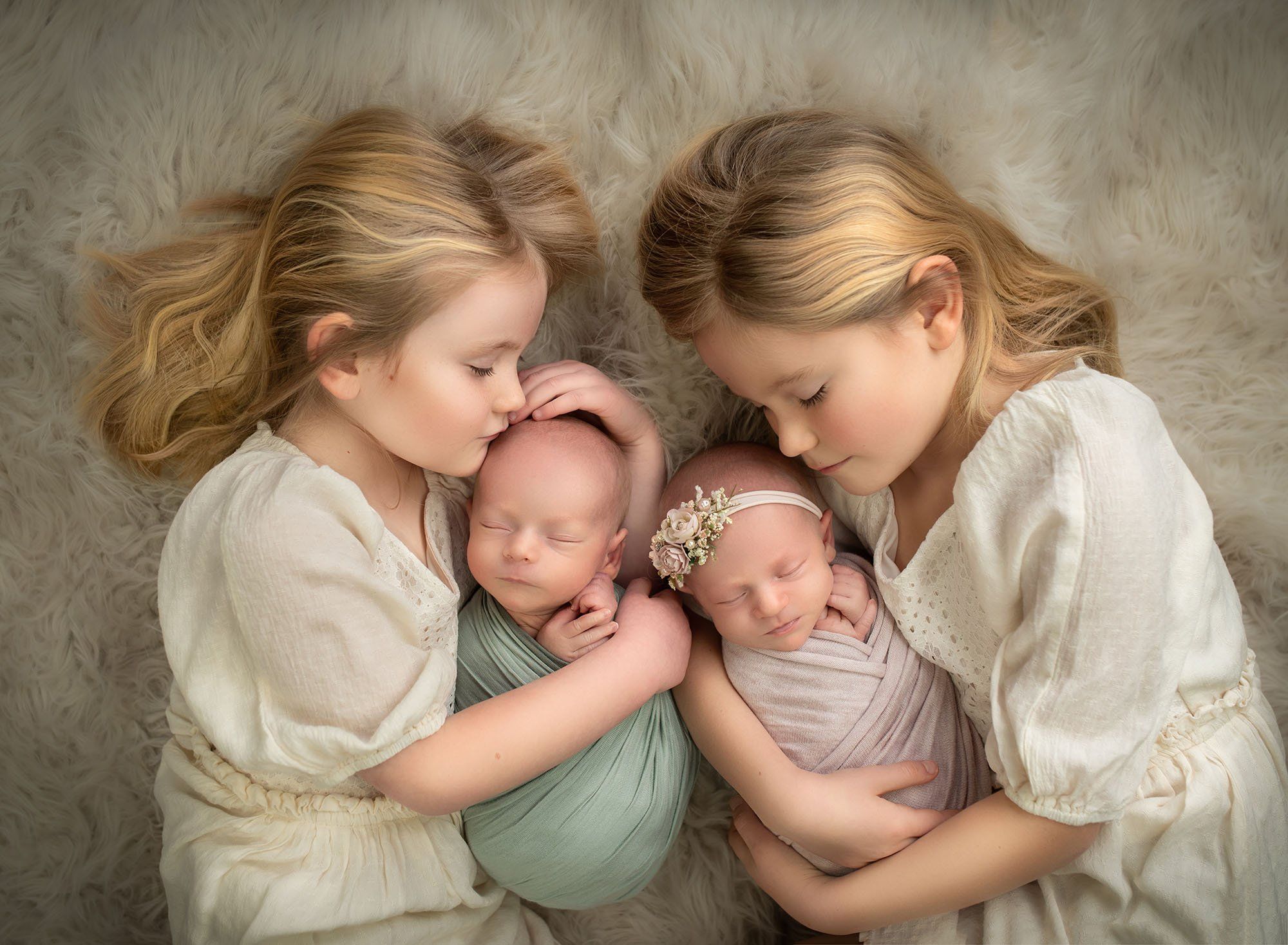Best Newborn Photographer CT blonde little girl sisters holding their newborn twin siblings while lying on a oatmeal color fur rug