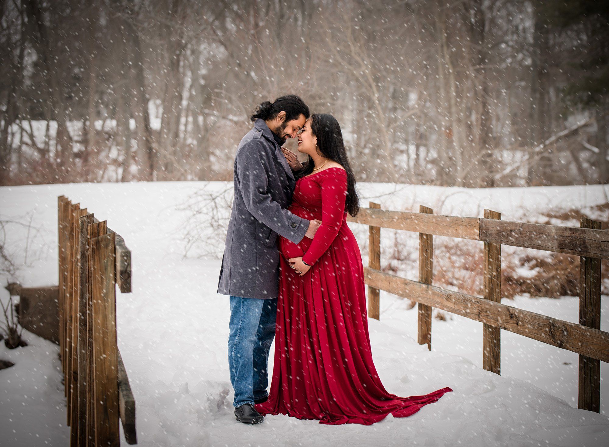 couples maternity photo with red maternity dress on a snowy bridge with falling snow