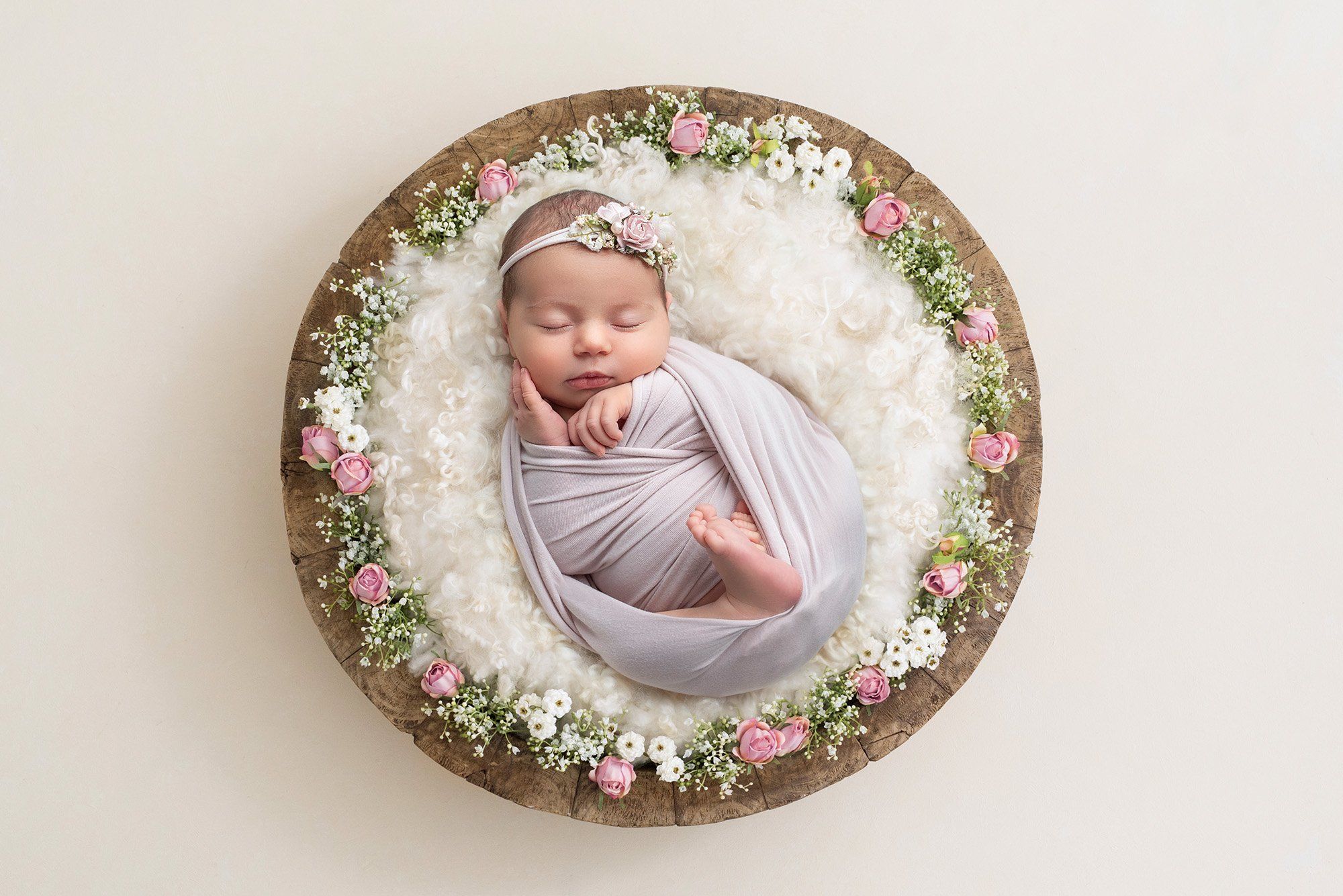 newborn baby girl asleep in wooden bowl atop a fluffy white blanket surrounded by pink roses and baby's breath