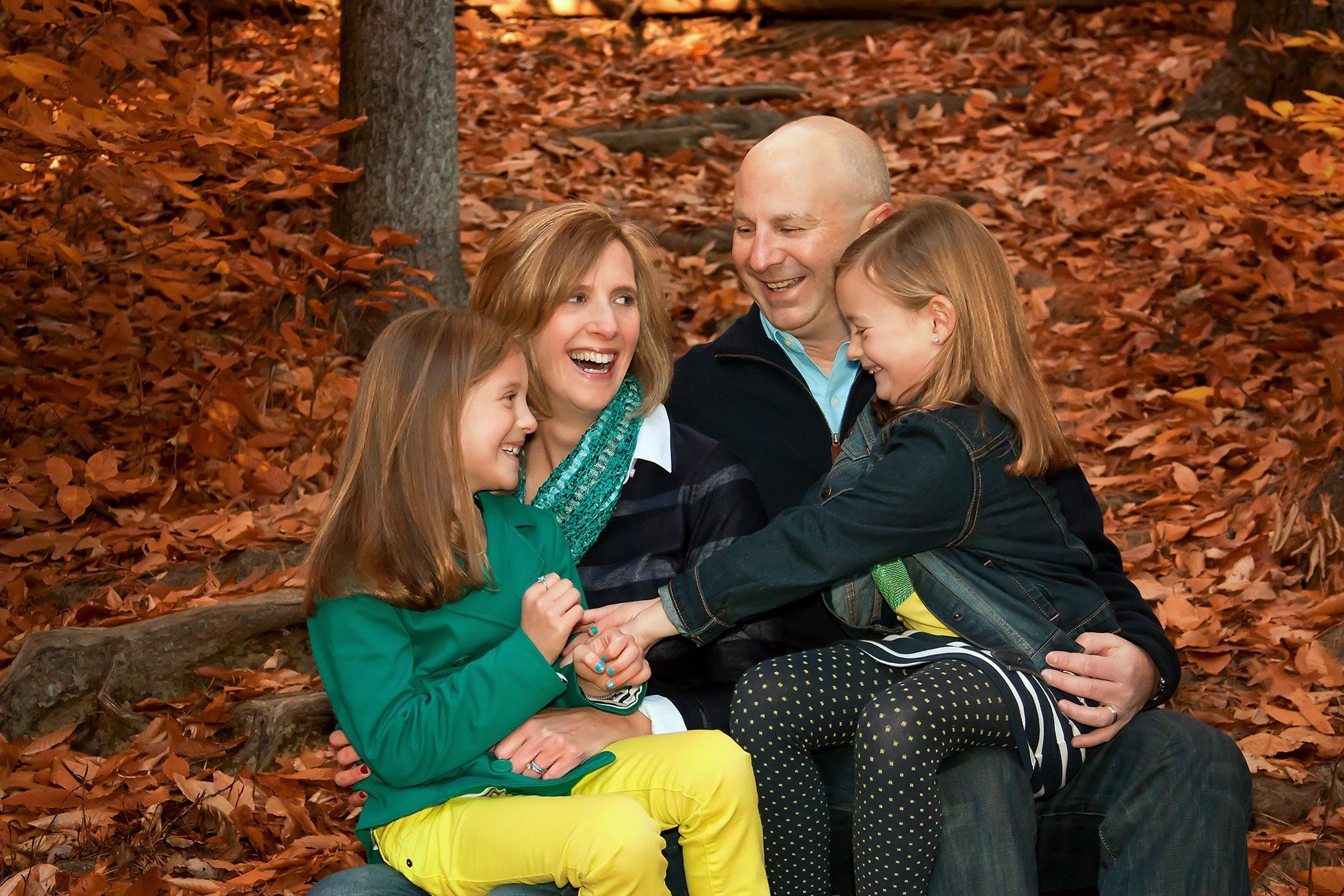 Mom, Dad and twin 7 year old girls sitting in orange fall leaves and laughing together