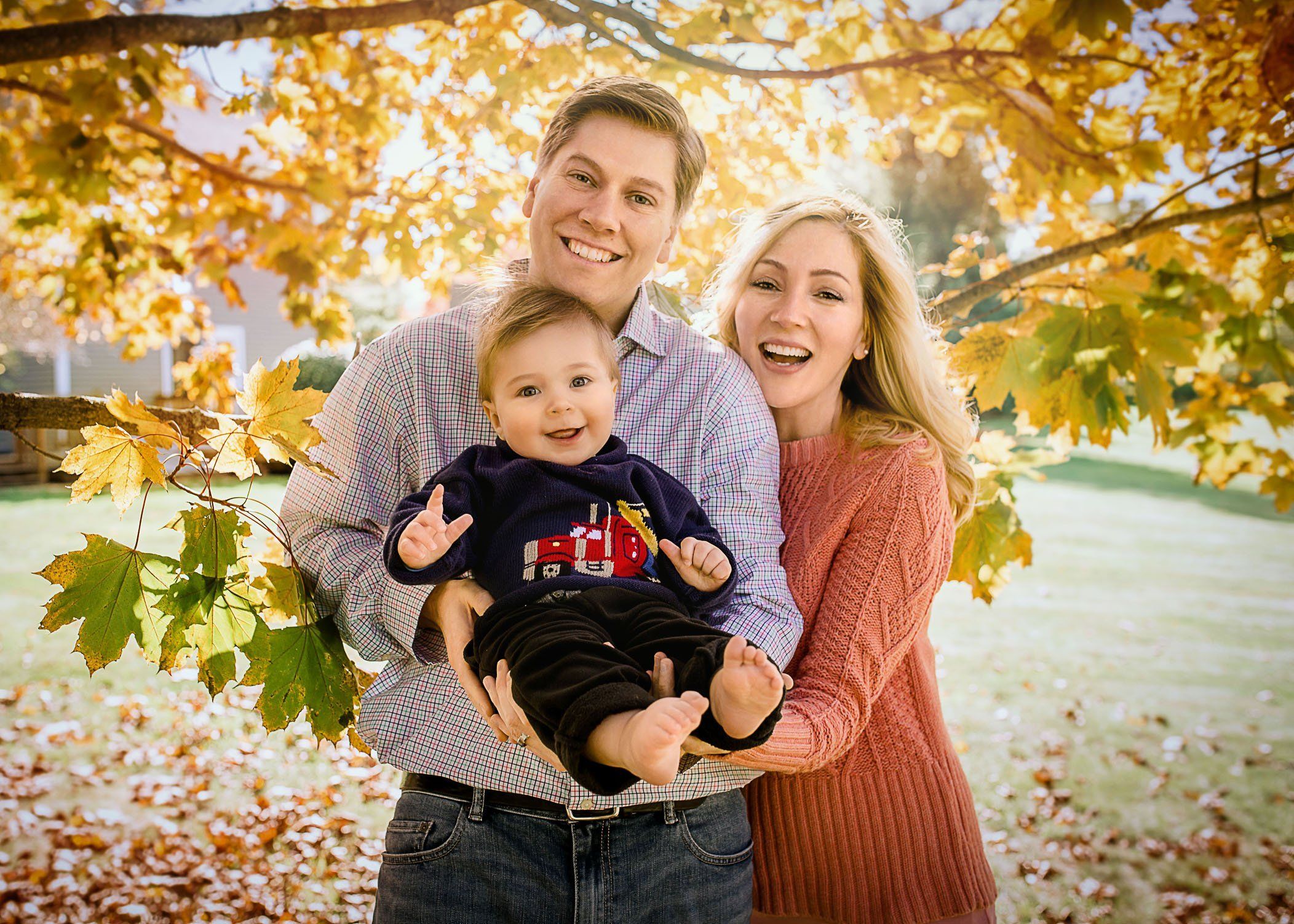 Mom Dad and baby boy smiling in yellow fall leaves outside One Big Happy Photo