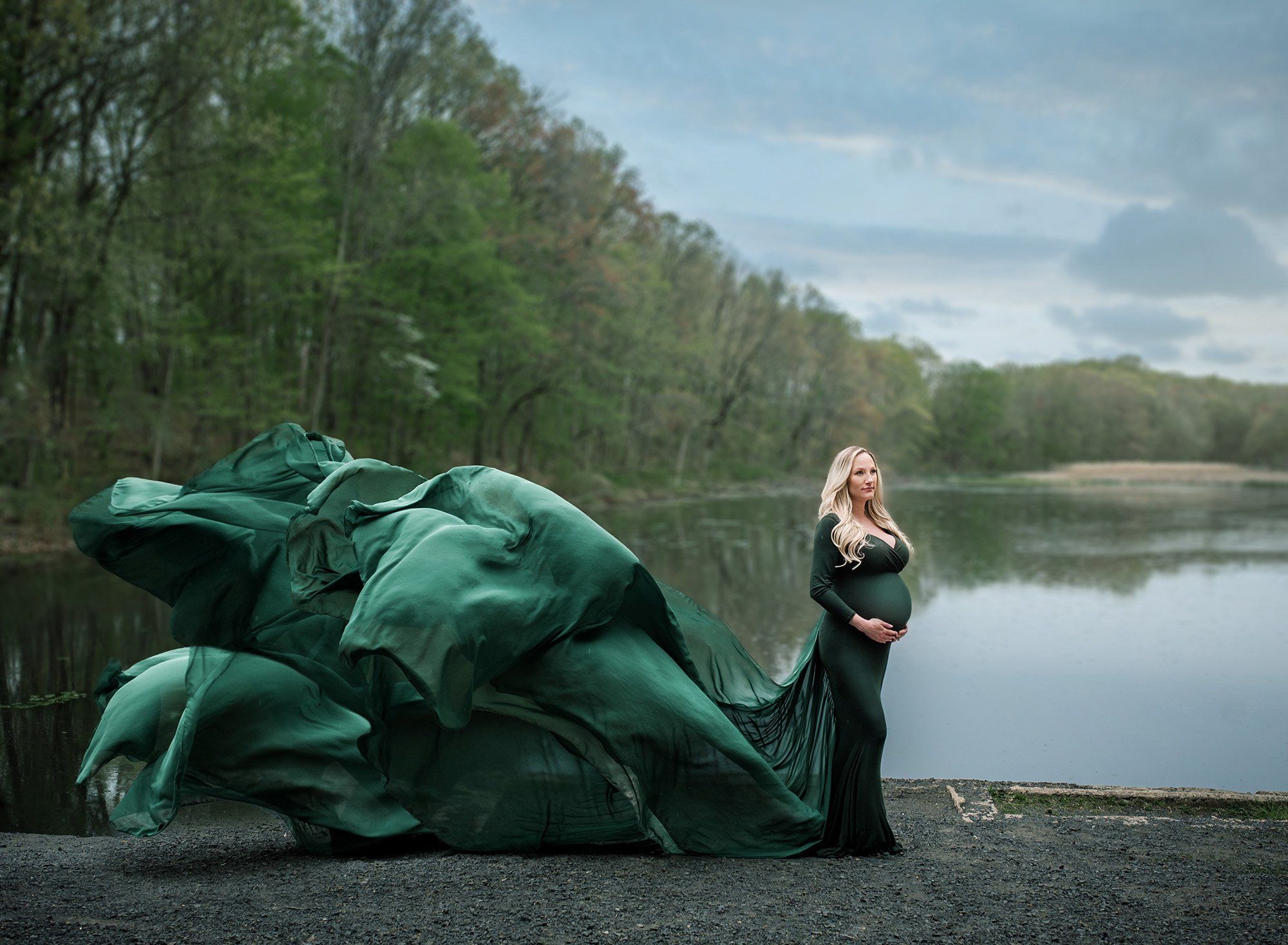 blonde pregnant woman dressed in green flowing maternity gown in front of lake on a cloudy day