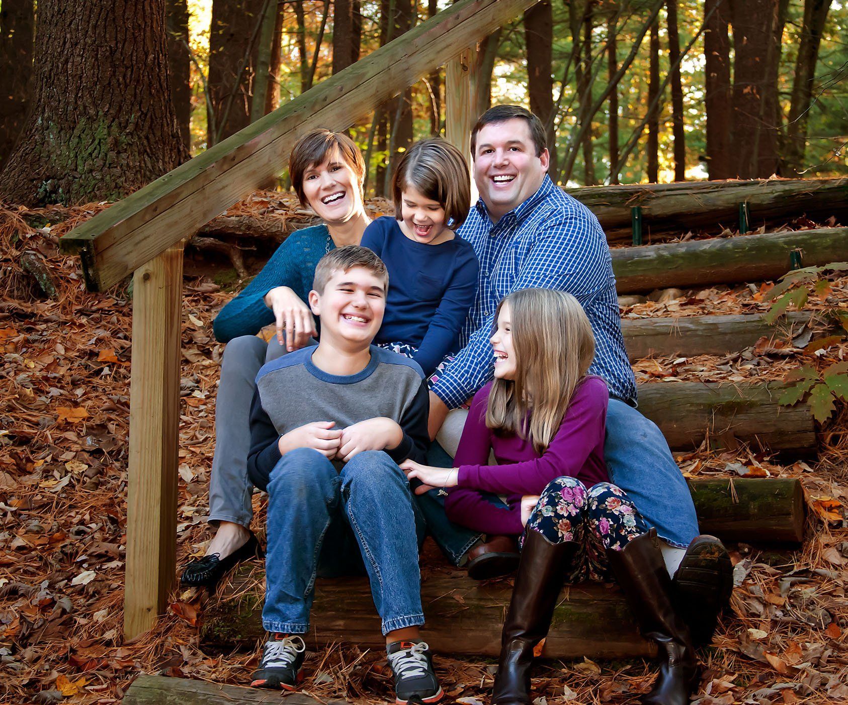Mom, Dad and 3 kids laughing and tickling each other outside on the stairs in the woods with leaves on the ground