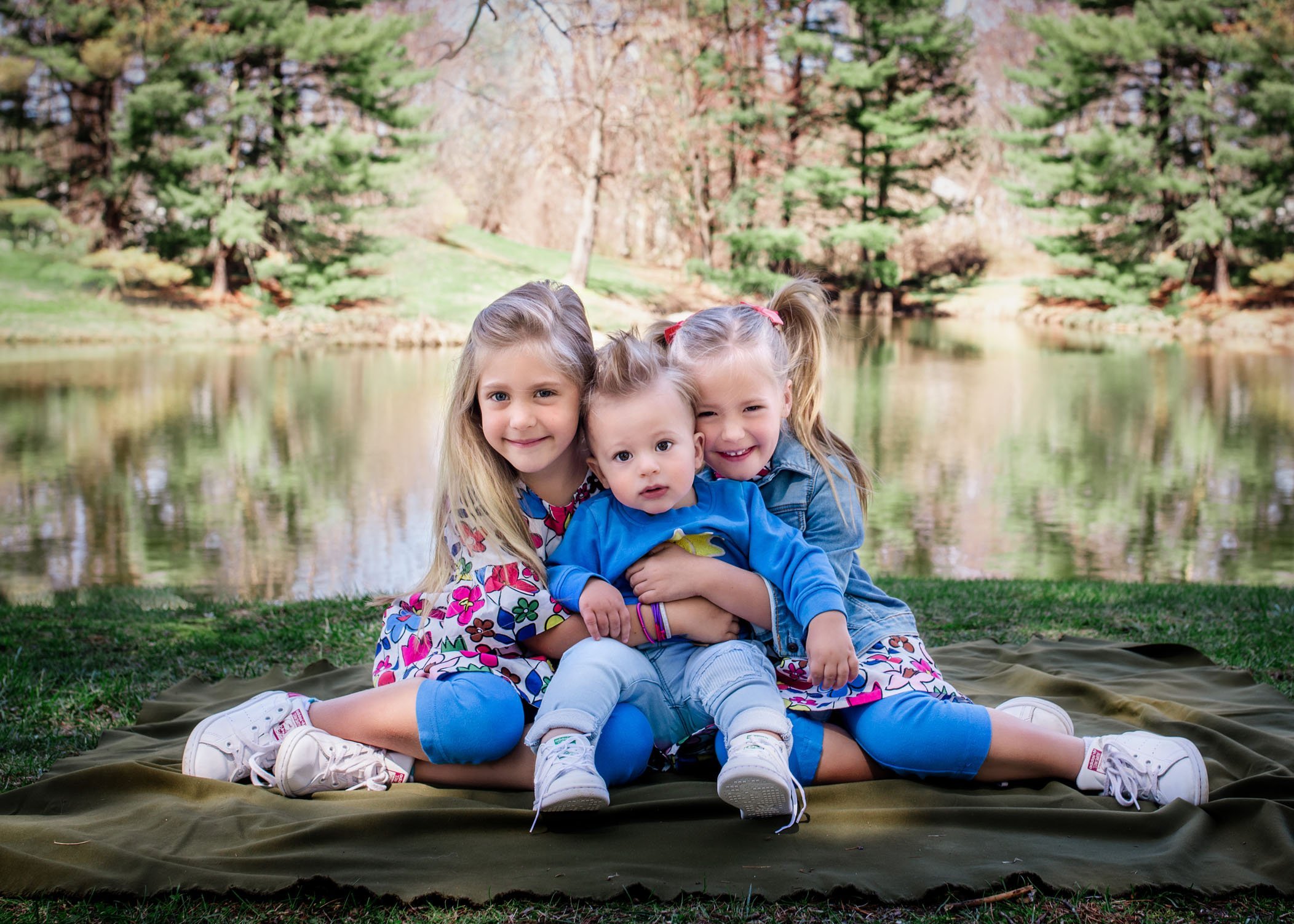 two big sisters and their 1 year old baby brother in park in front of pond