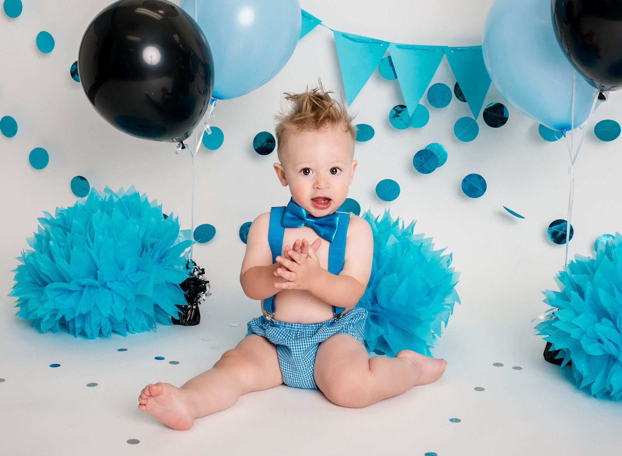 year old boy sitting on white, blue and black decorated backdrop in blue suspenders