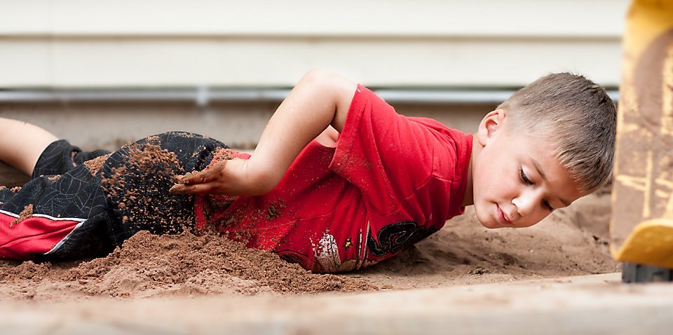 little boy lying on the ground in a red shirt and black shorts, digging a hole with his hands in the dirt