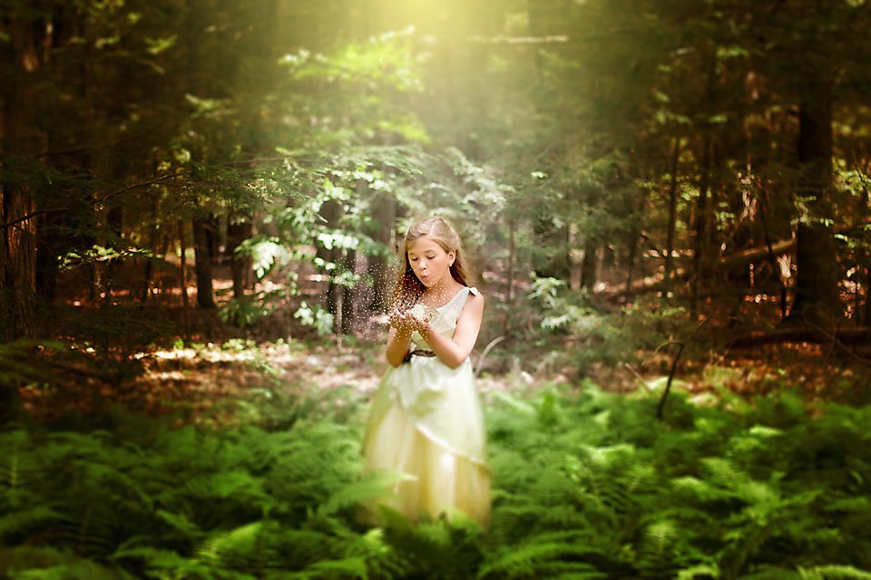 little girl standing in a green forest with sun light shining down on her as she blows a handful of fairy dust 