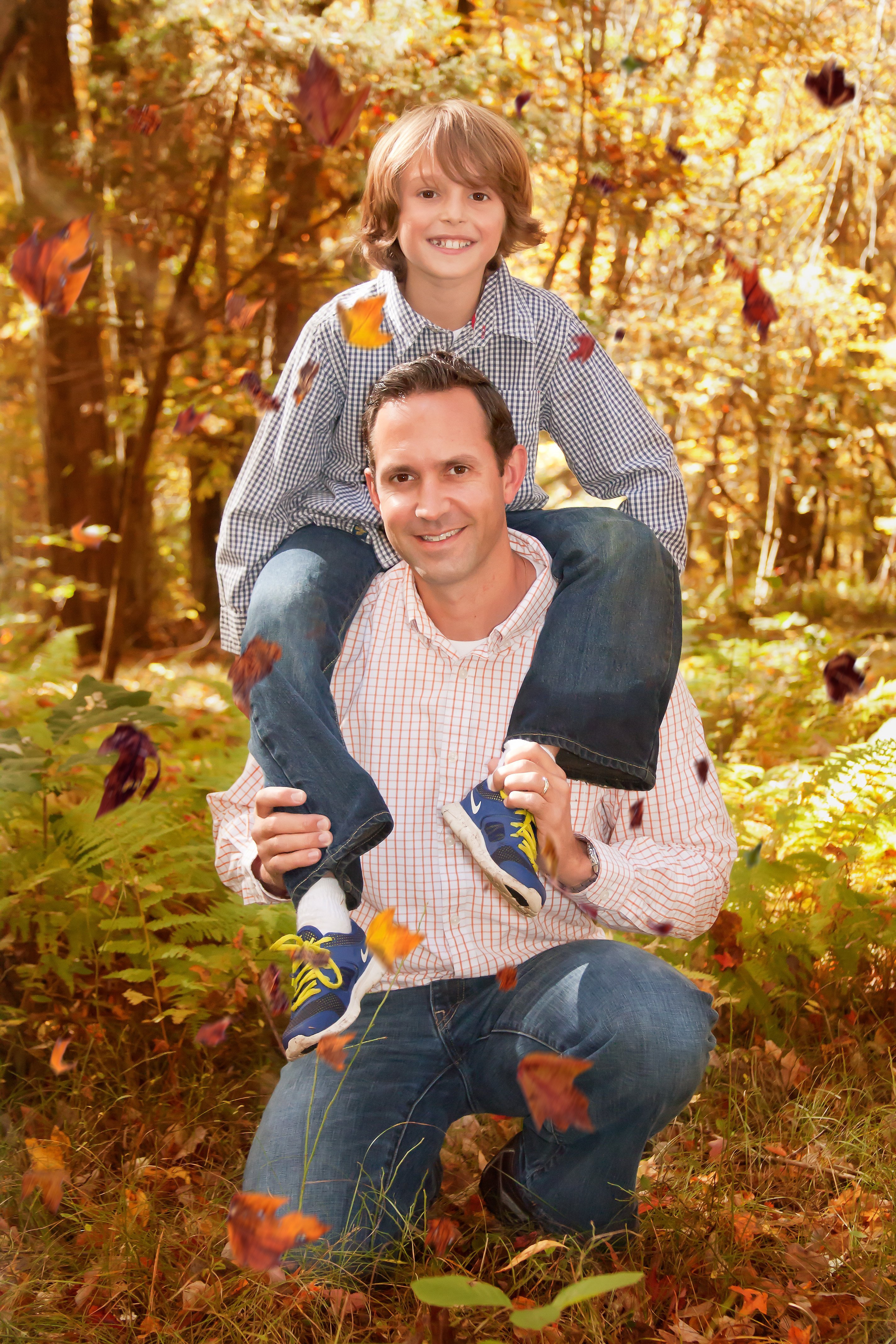 Dad with young son on his shoulders in the woods with fall leaves swirling in the wind