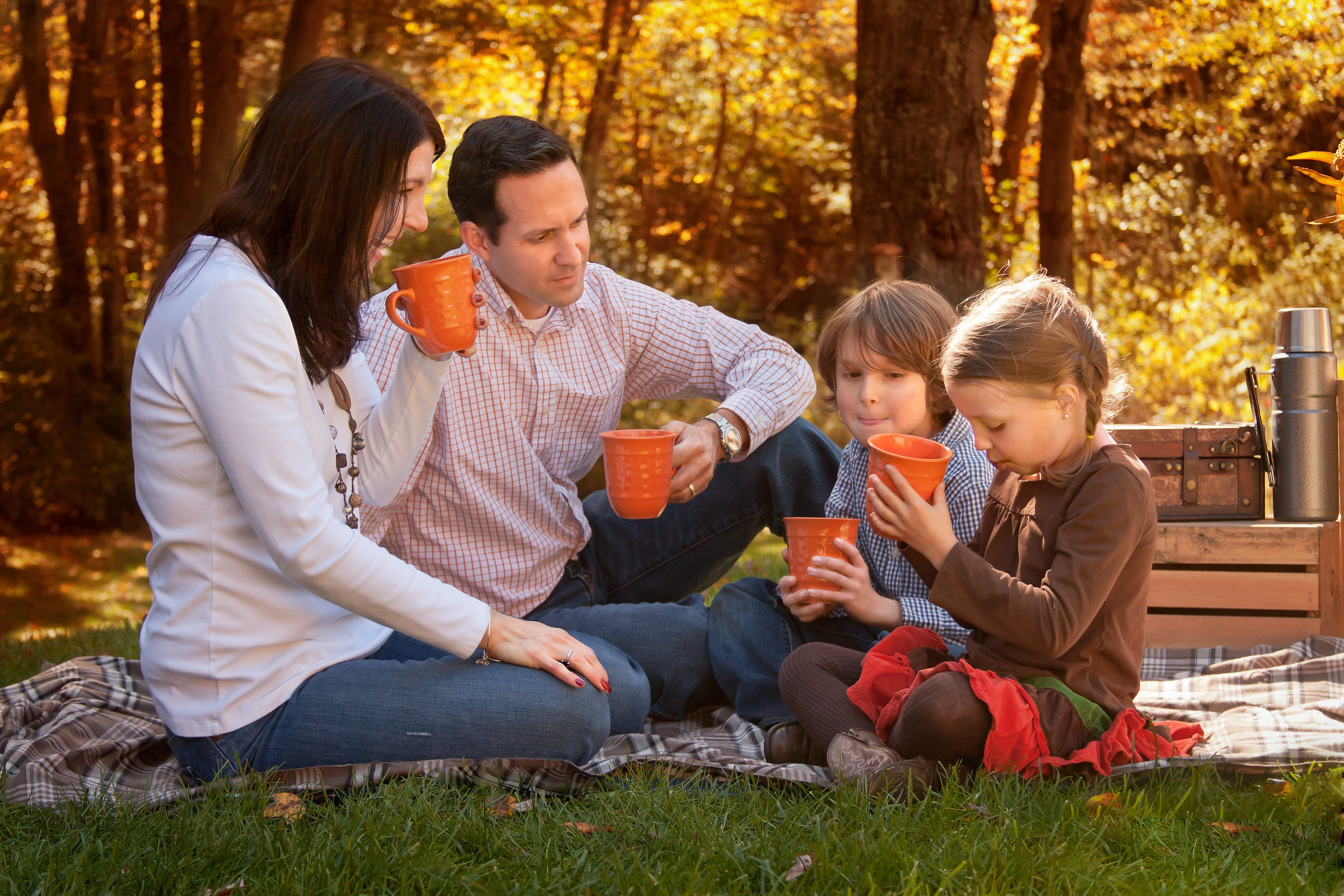 Mom, Dad and two kids enjoying hot cocoa in the fall sitting on a blanket with orange mugs