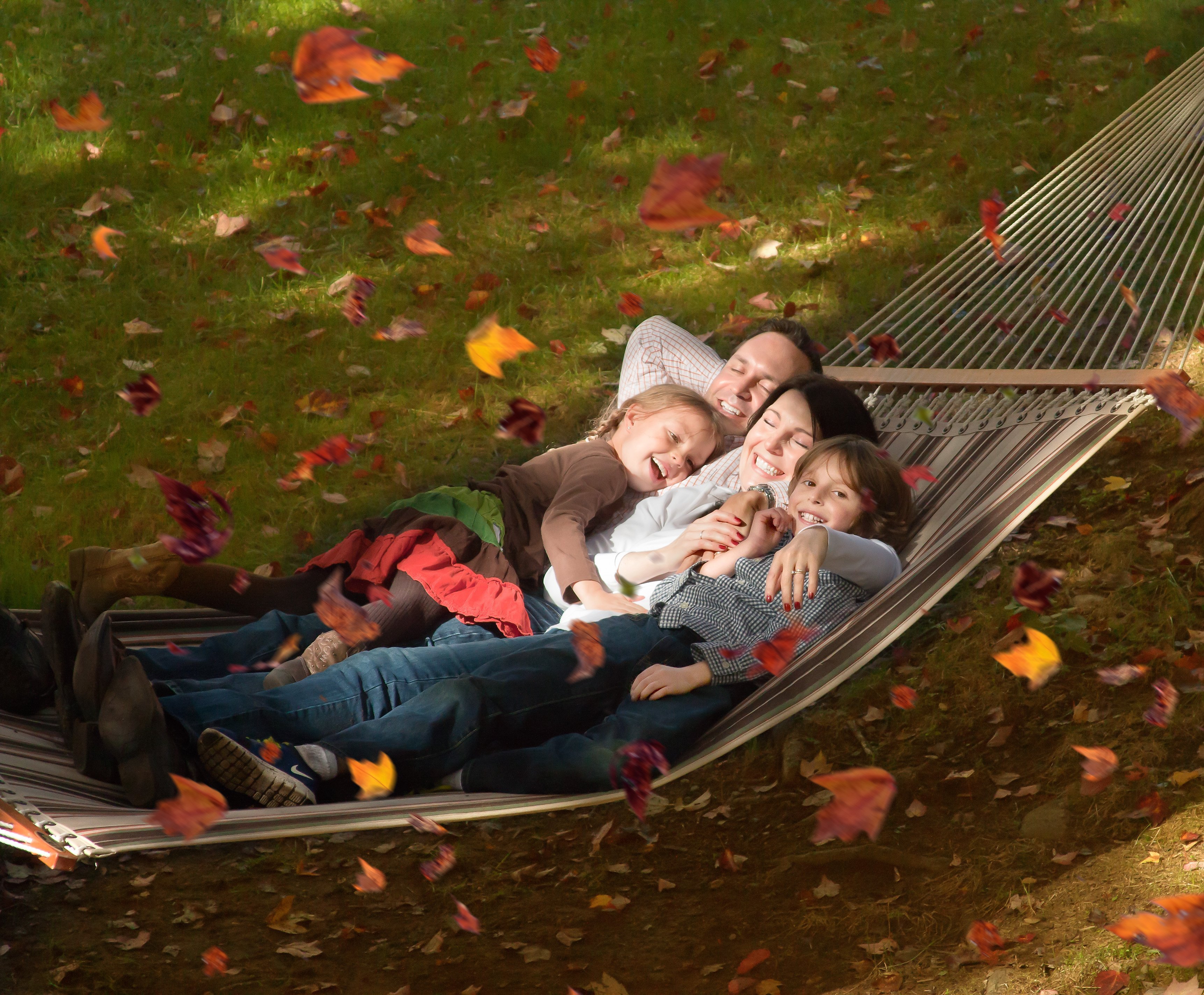 Mom, Dad and 2 kids laughing while swinging in a hammock with fall leaves swirling in the wind