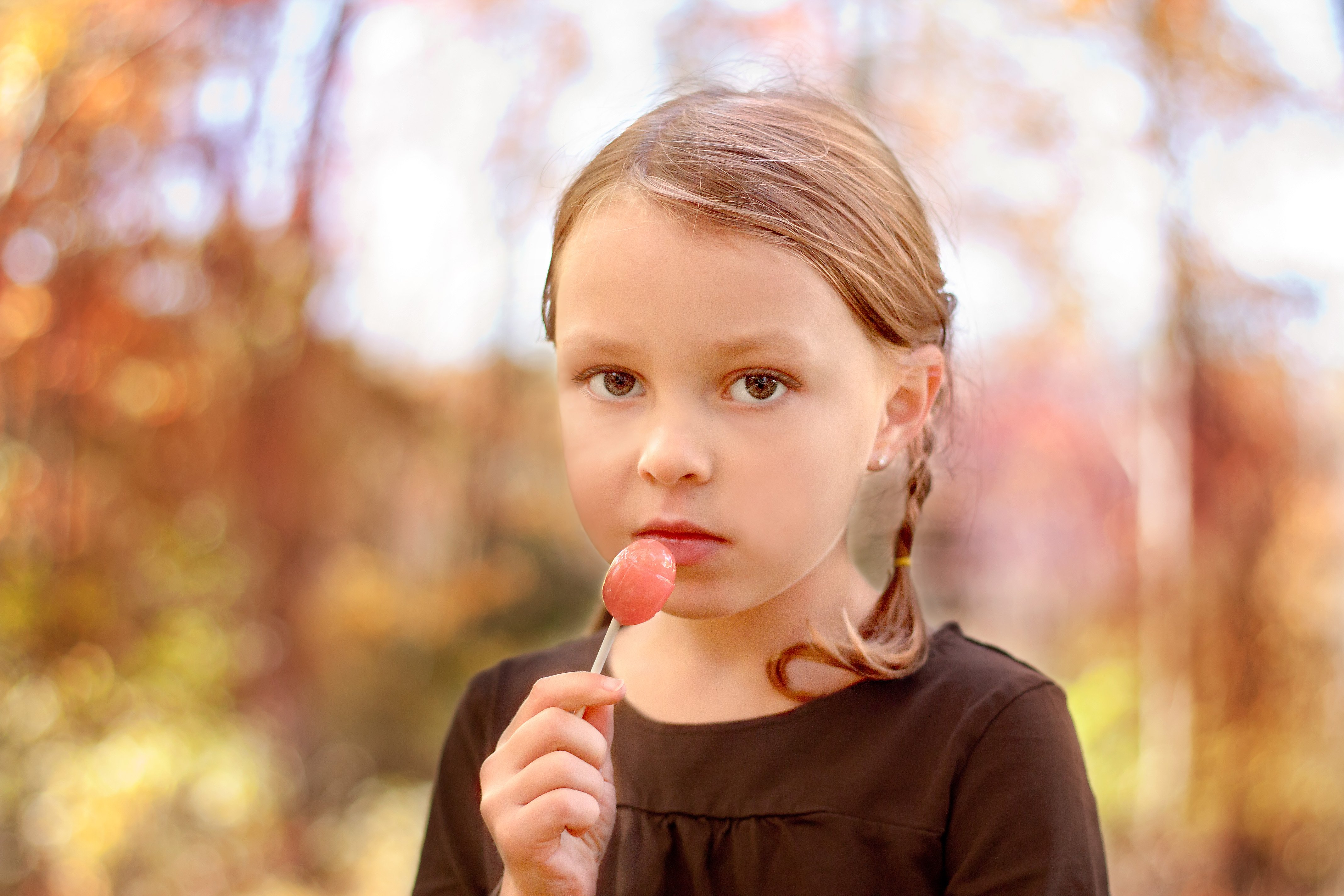 young girl with a serious angelic look eating a pink lollipop with fall colors behind her