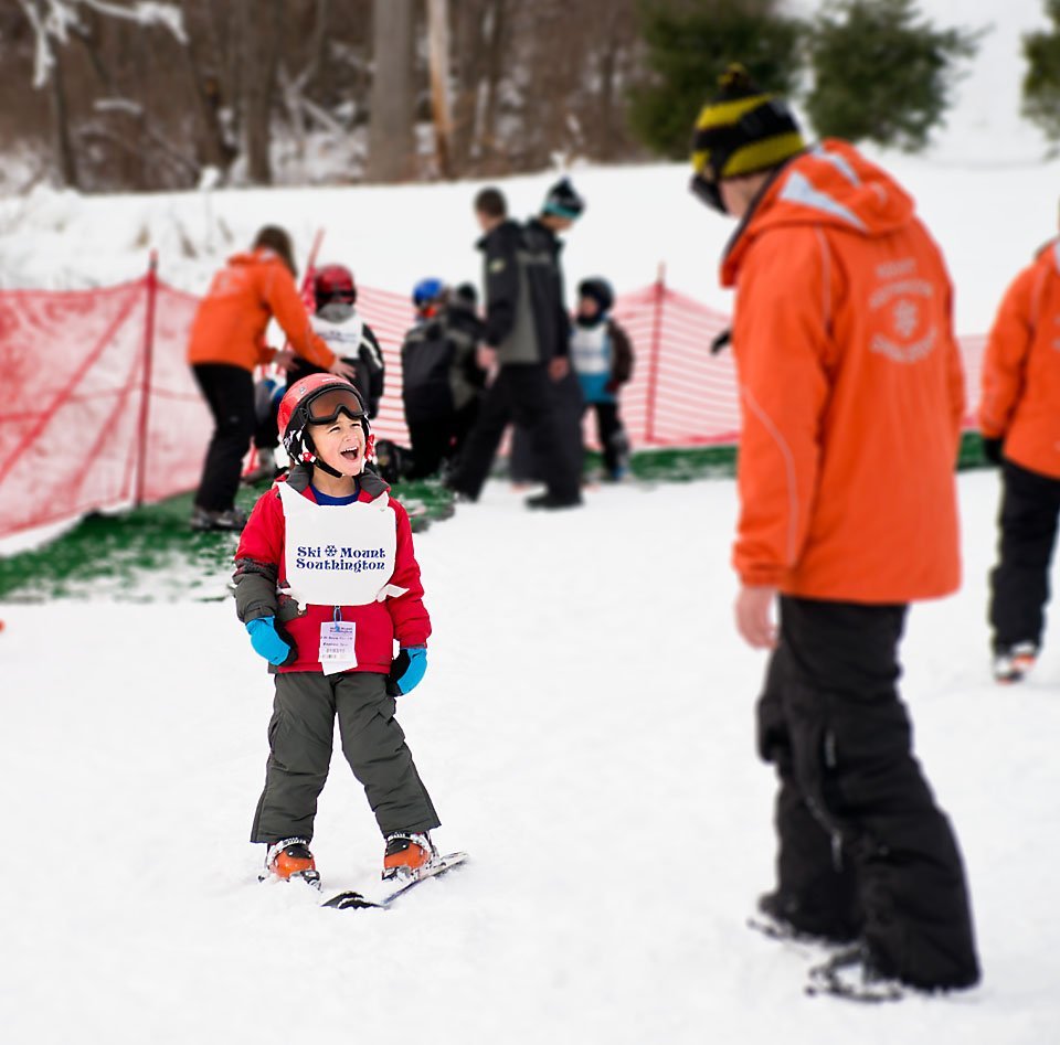 5 year old boy in bright red winter ski clothes laughing with his skiing instructor