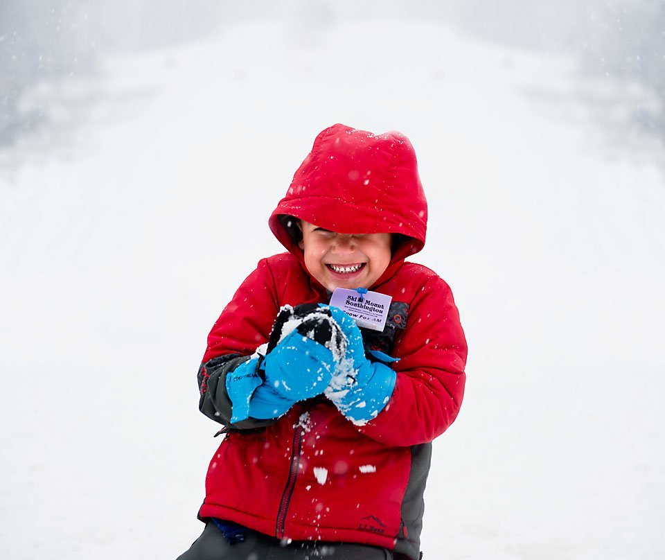 5 year old boy in a read winter coat smiling while he makes a snowball in the falling snow