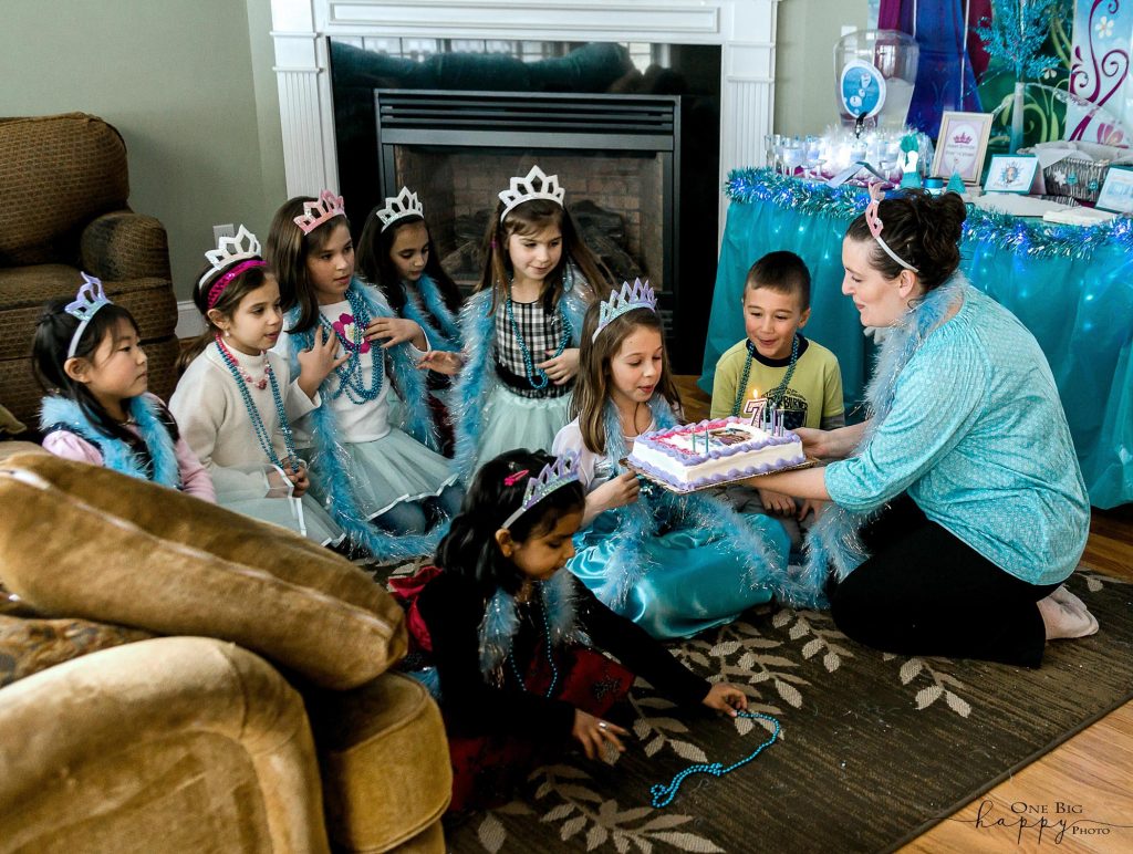 little girls dressed in blue as princesses surround one girl as she blows out the candles on her 7 year old birthday cake