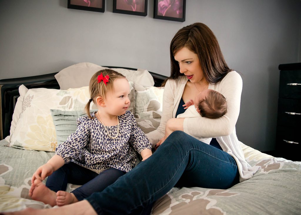 toddler sits with mom and newborn sister on moms bed