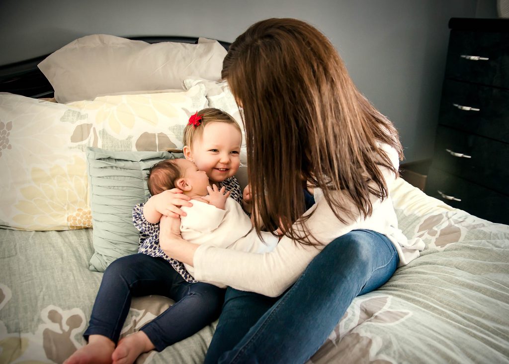 toddler girl smiles while holding newborn sister with mom's help