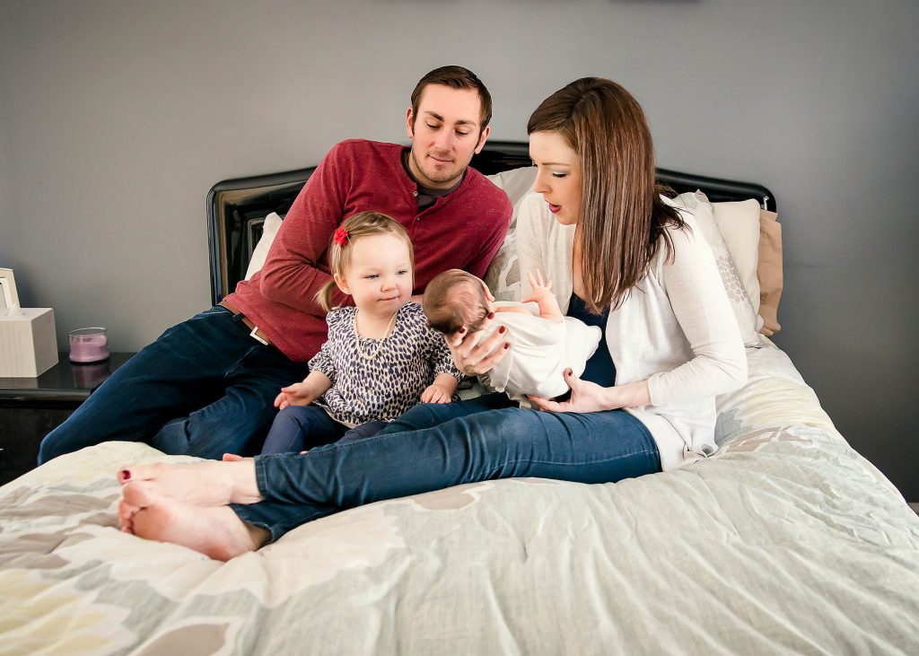 Mom, Dad and two young daughters sit on parents bed