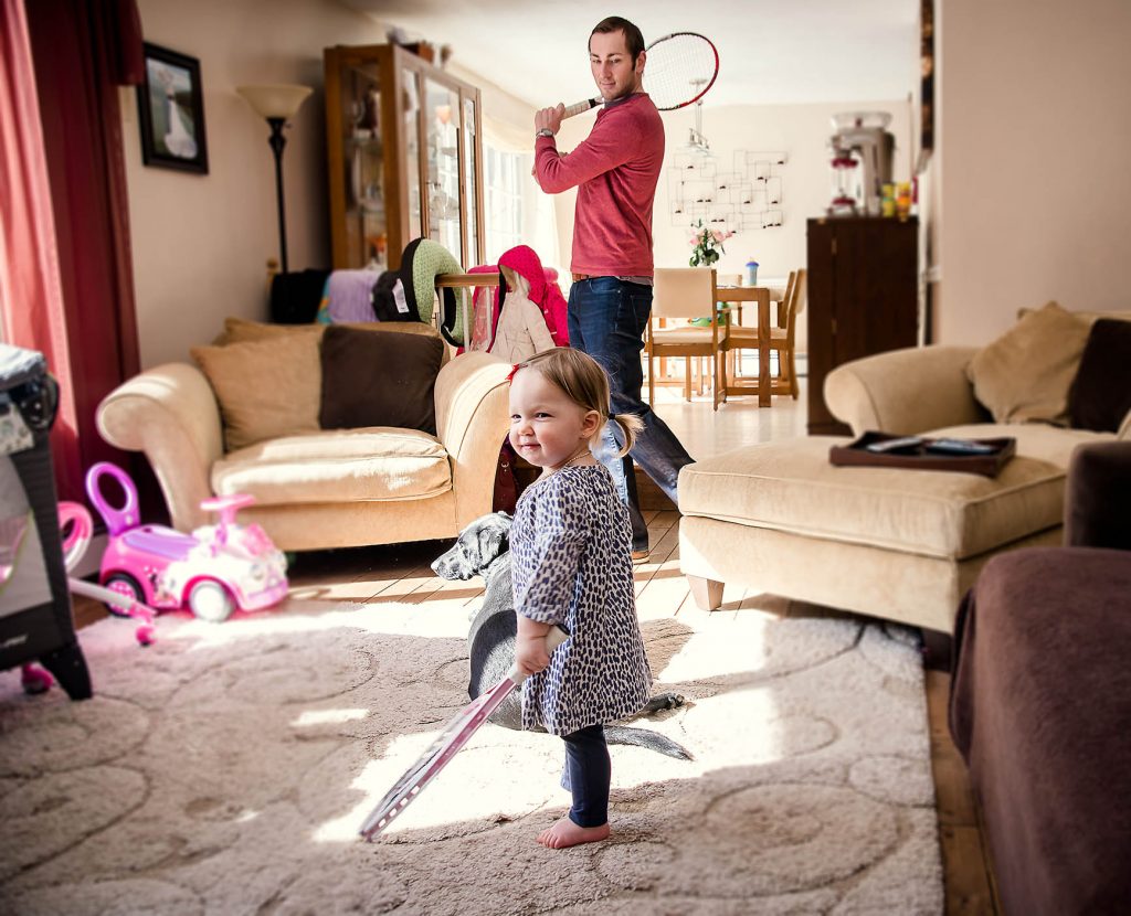 toddler girl holds a tennis racket while Dad practices his swing in the background