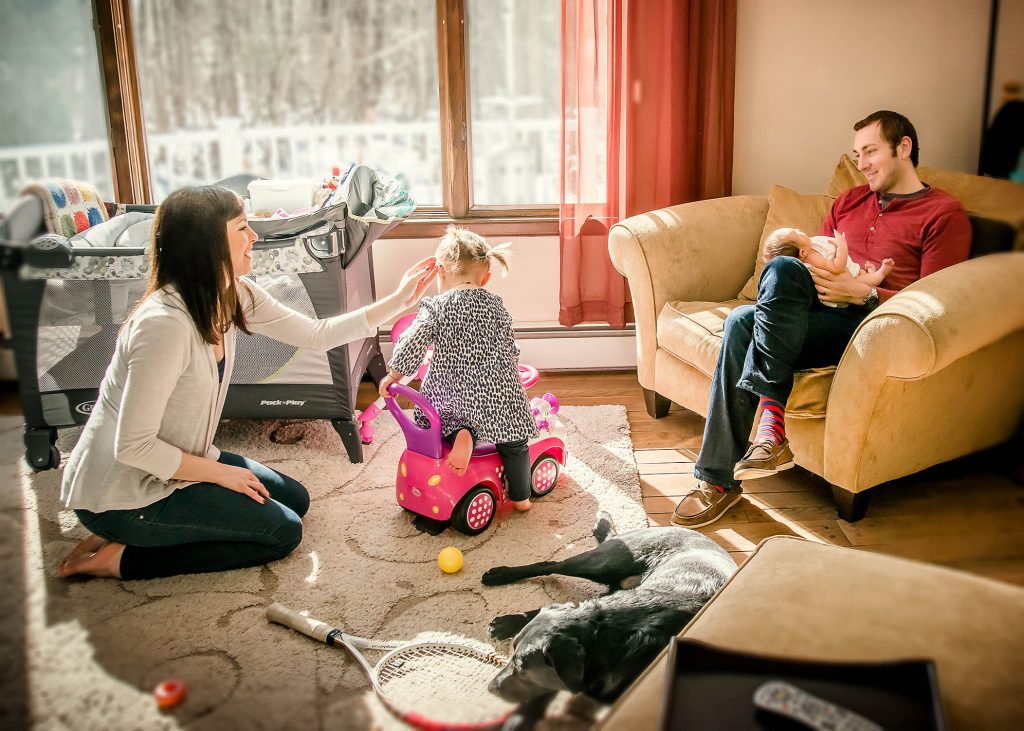 parents look on while toddler plays in the living room with Dad holding newborn daughter