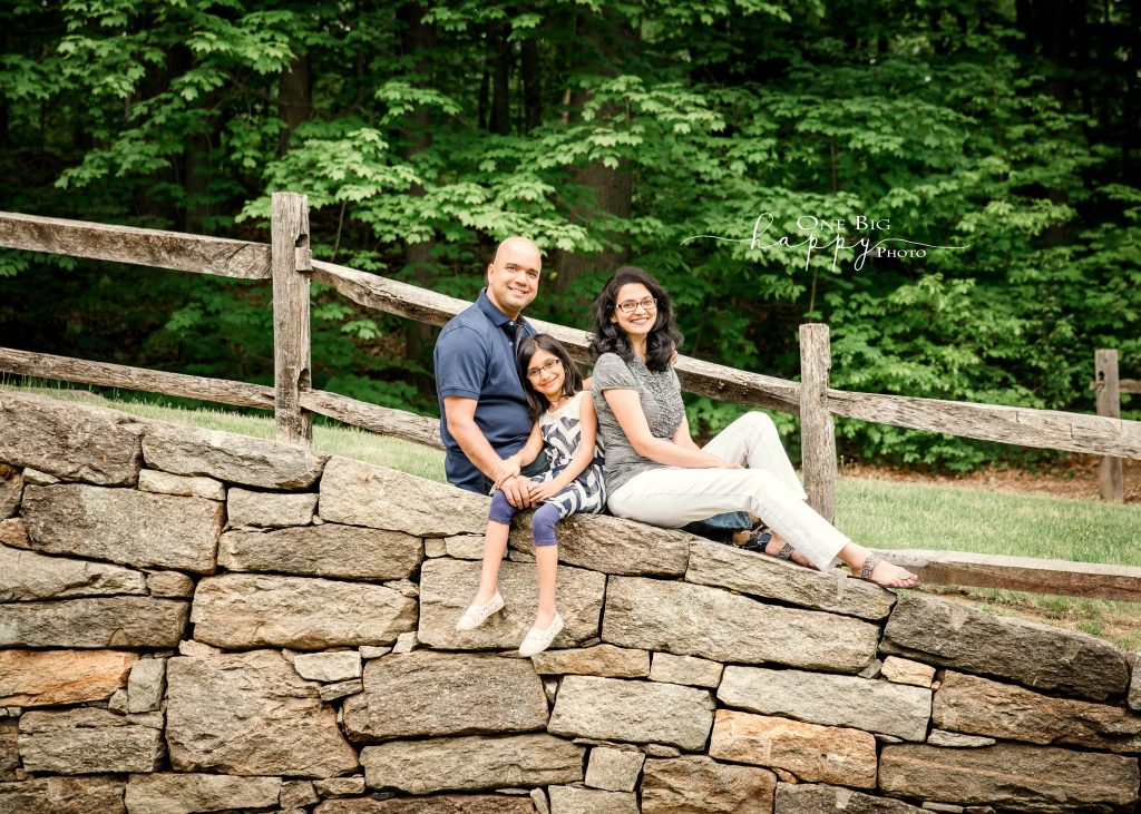 Dad, daughter, and Mom portrait on rock wall with green trees in background