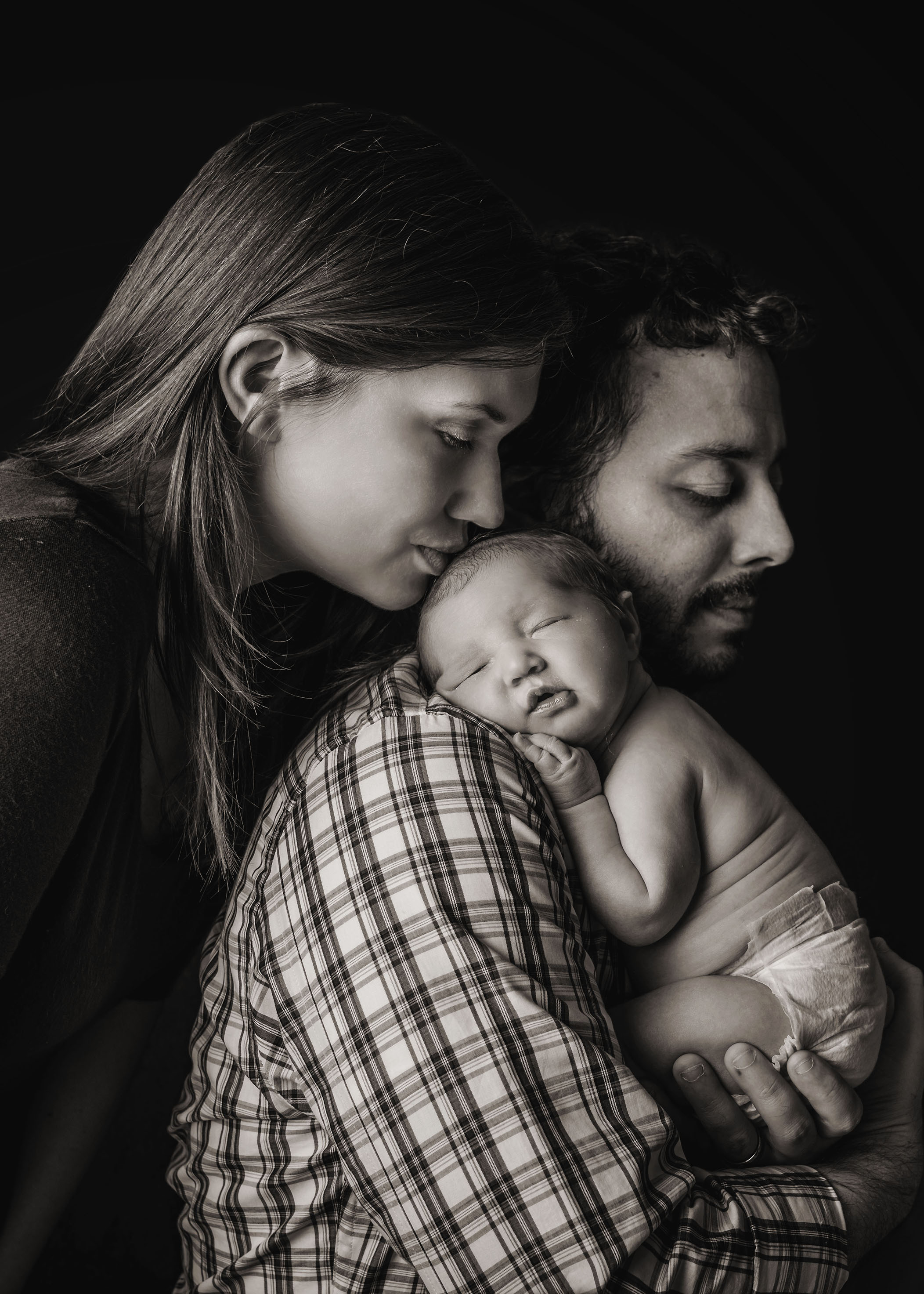 Dad holding newborn baby on his shoulder while mom kisses her head One Big Happy Photo