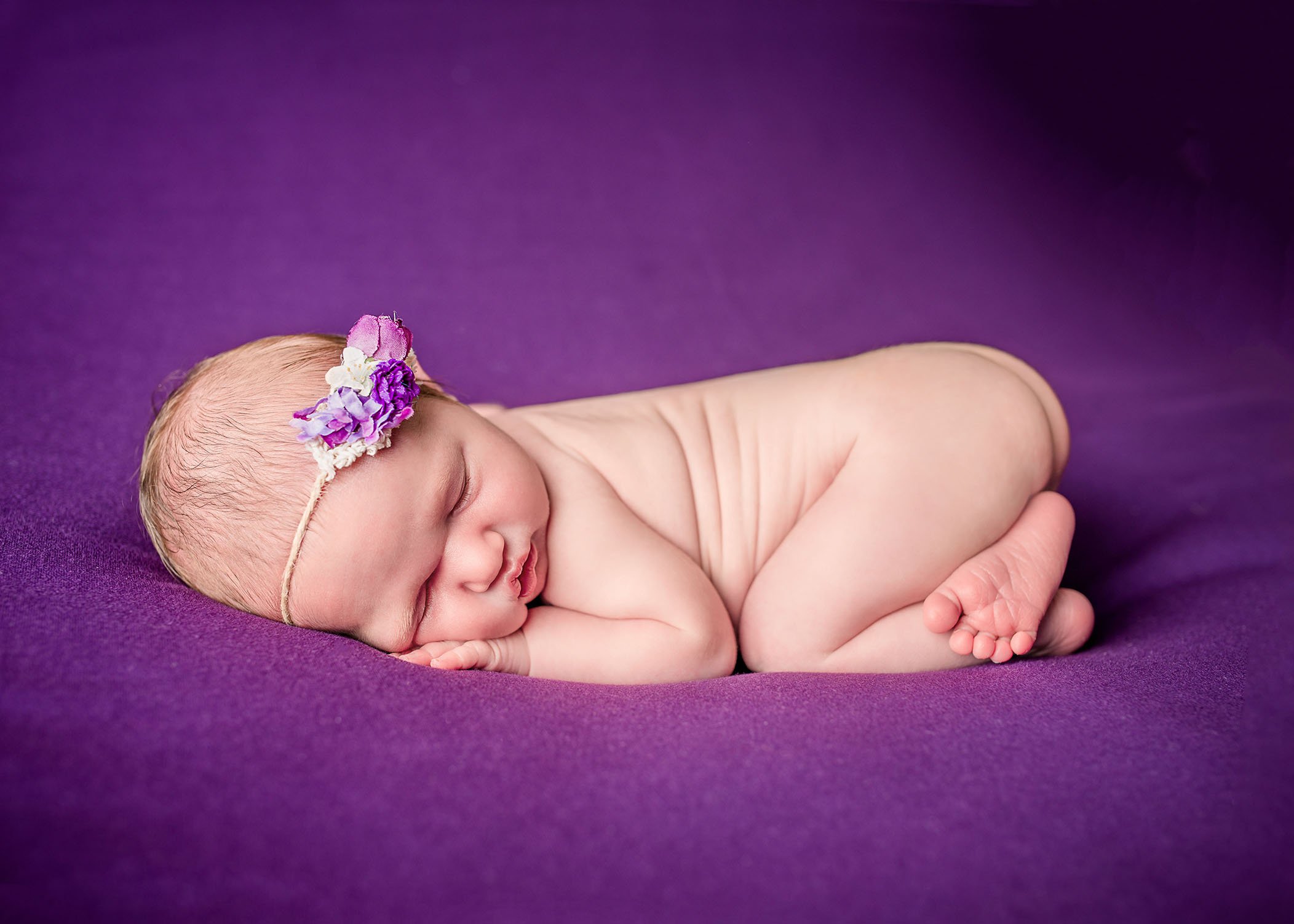 newborn baby girl sleeping on tummy on purple background with floral headband One Big Happy Photo