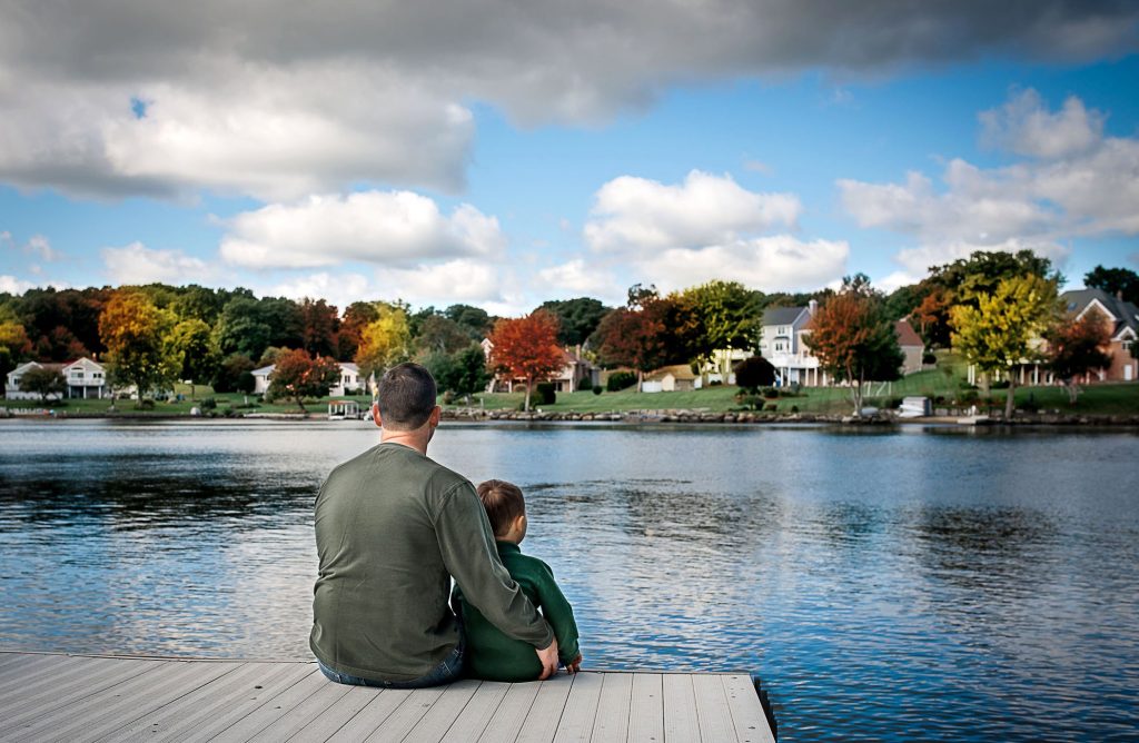Father and young son sitting on the dock of a lake looking at the fall foliage One Big Happy Photo