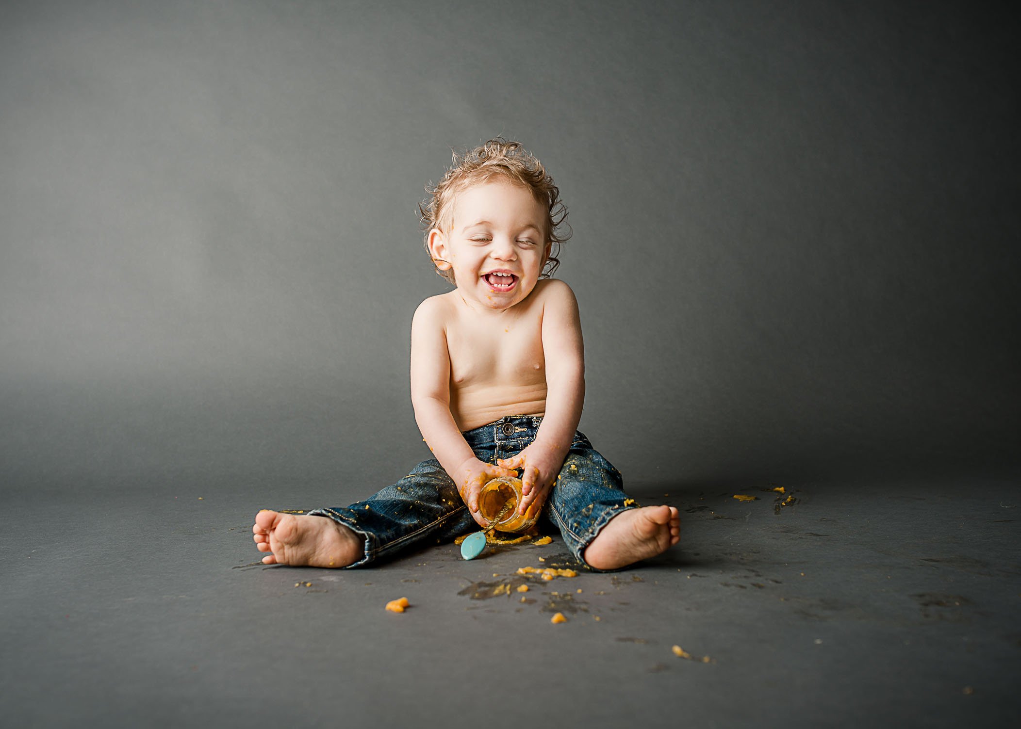 18 month old boy sitting on ground and messy from eating jar of food One Big Happy Photo