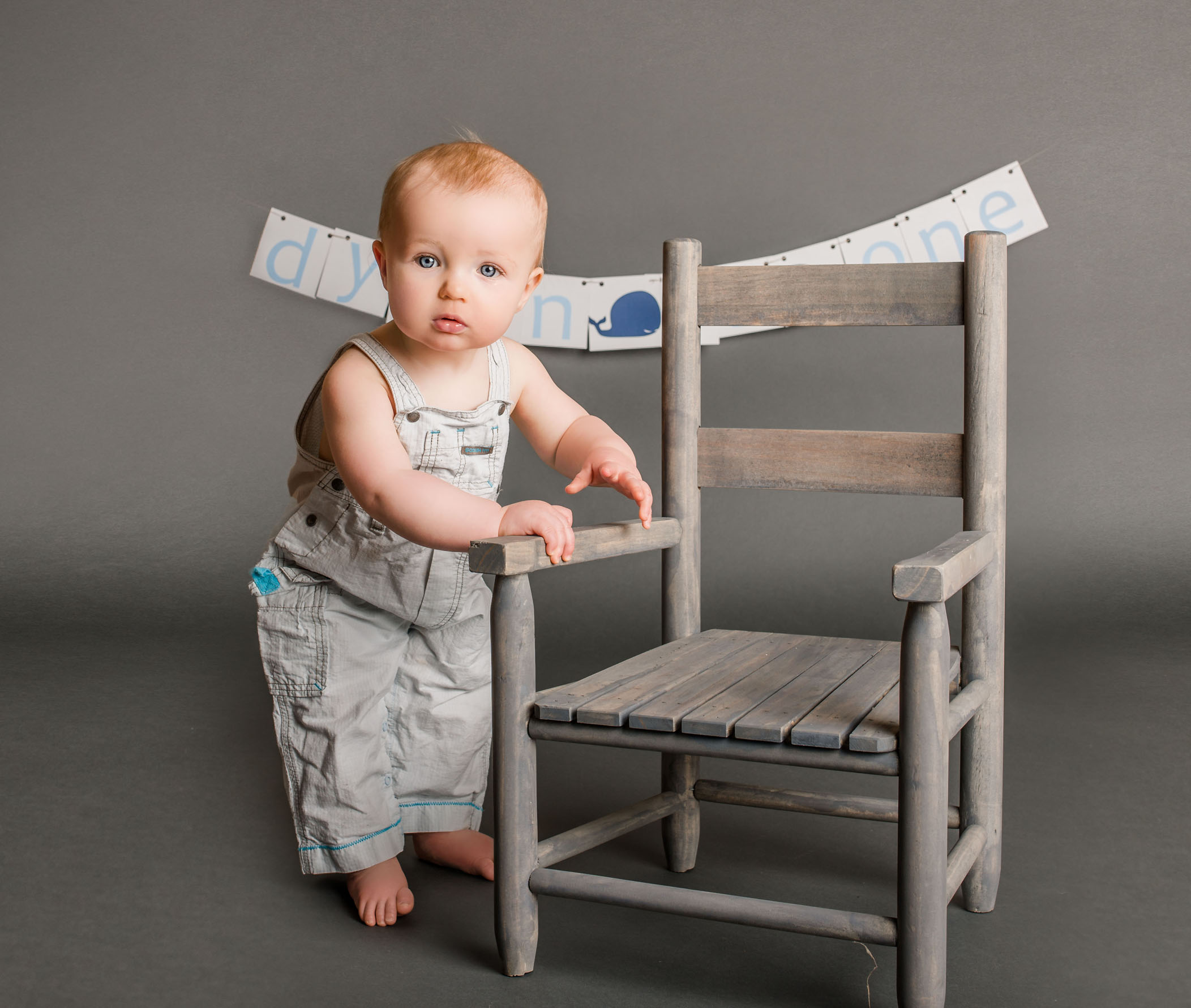one year old baby in overalls standing next to small chair One Big Happy Photo