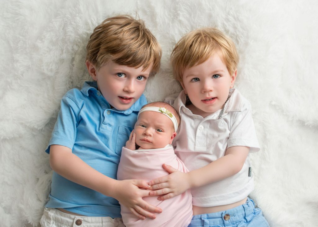 little brothers holding newborn baby sister on white rug