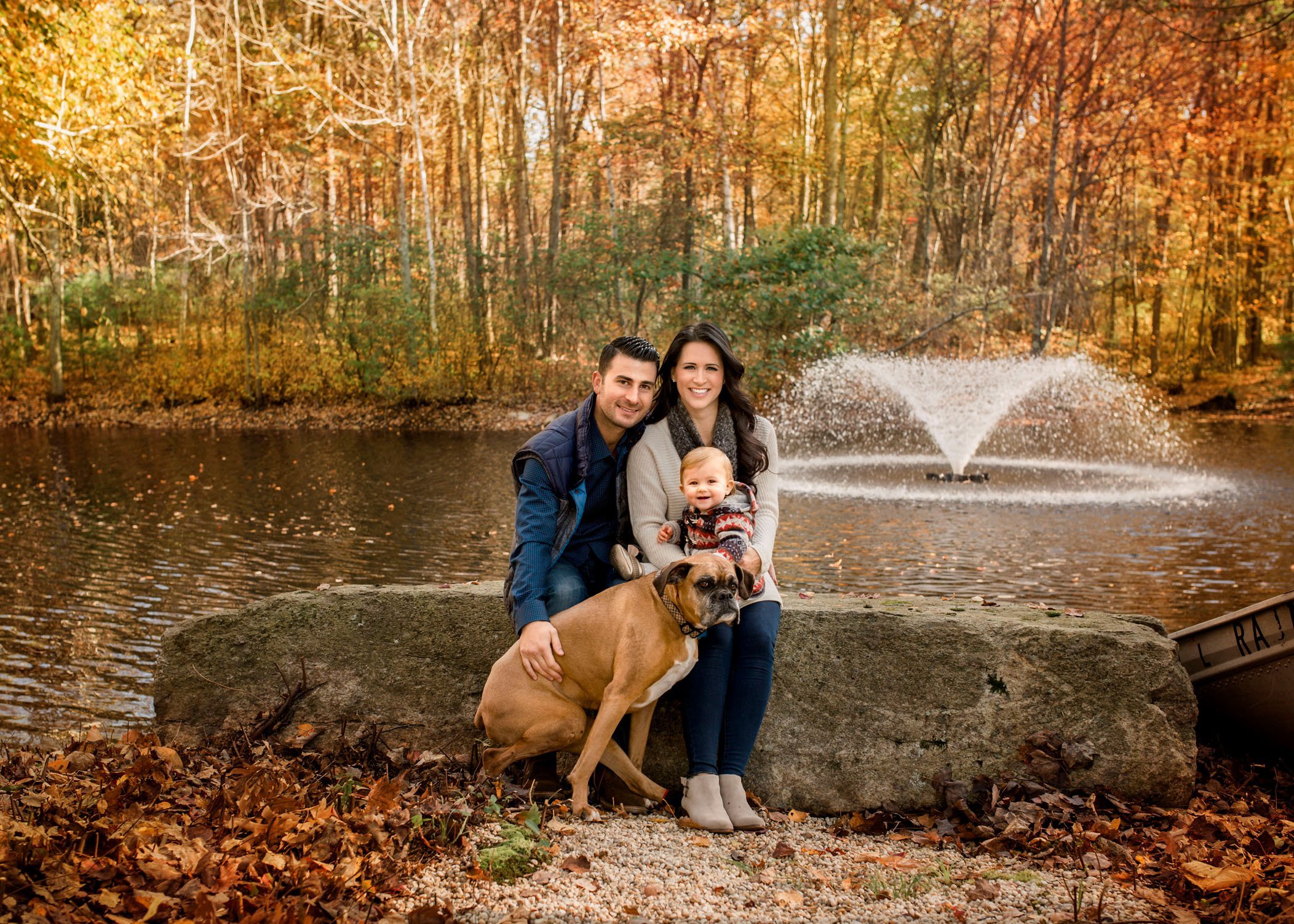 Mom Dad 1 year old daughter and dog sit on rock in front of pond outside in fall