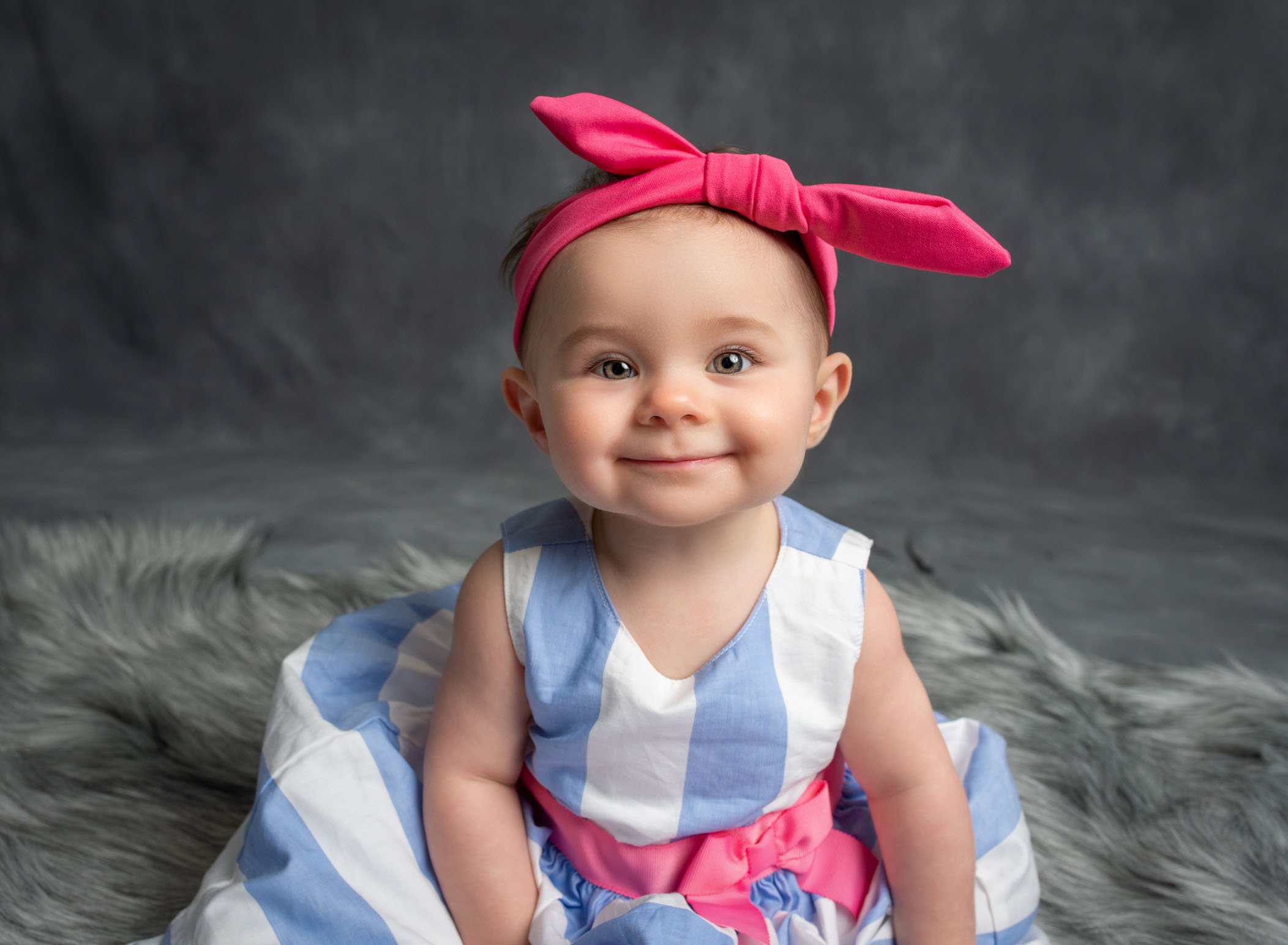 smiling baby girl sitting up on grey rug with large pink bow