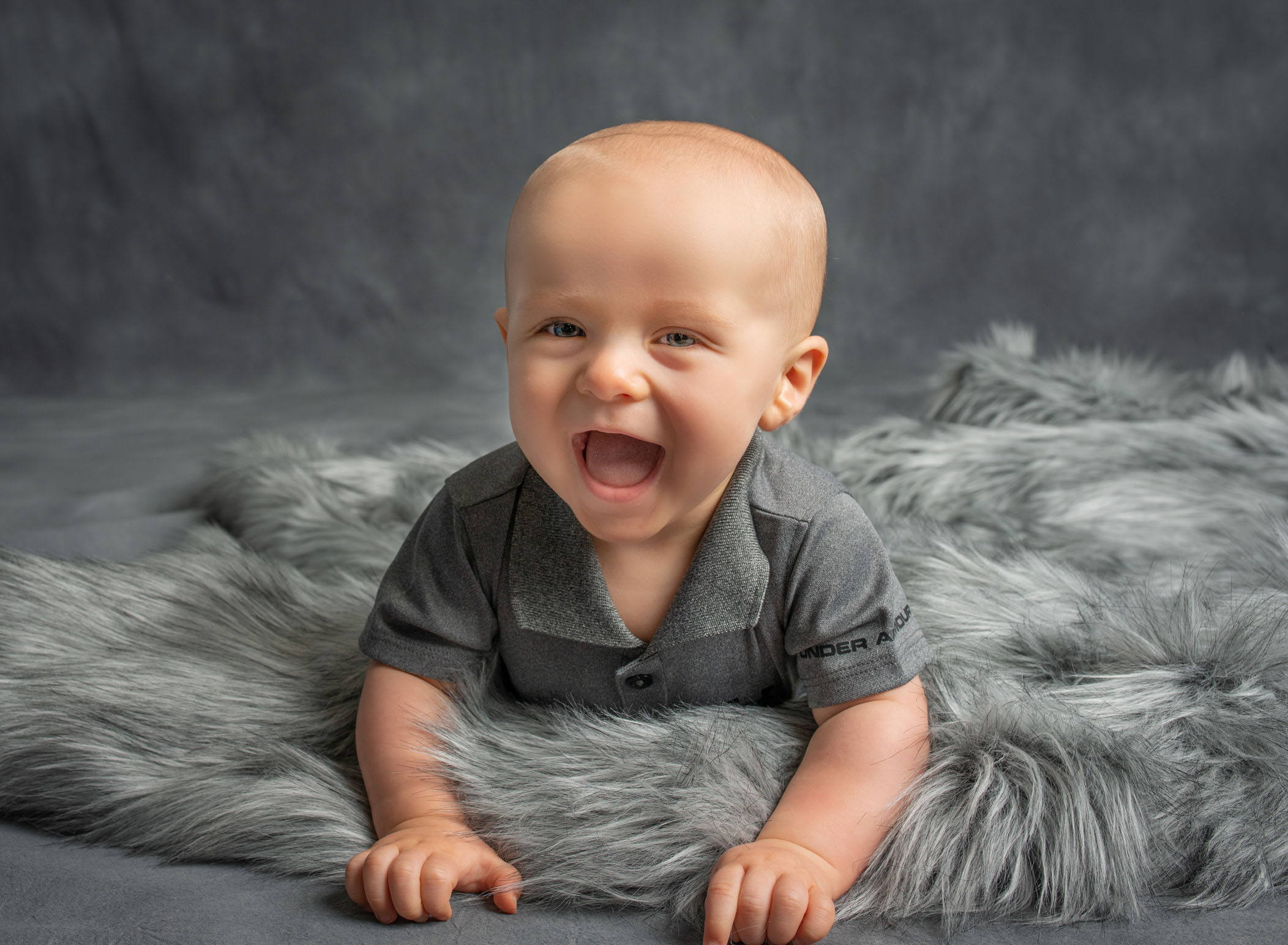 6 month old boy smiling wide on his tummy on grey fur blanket