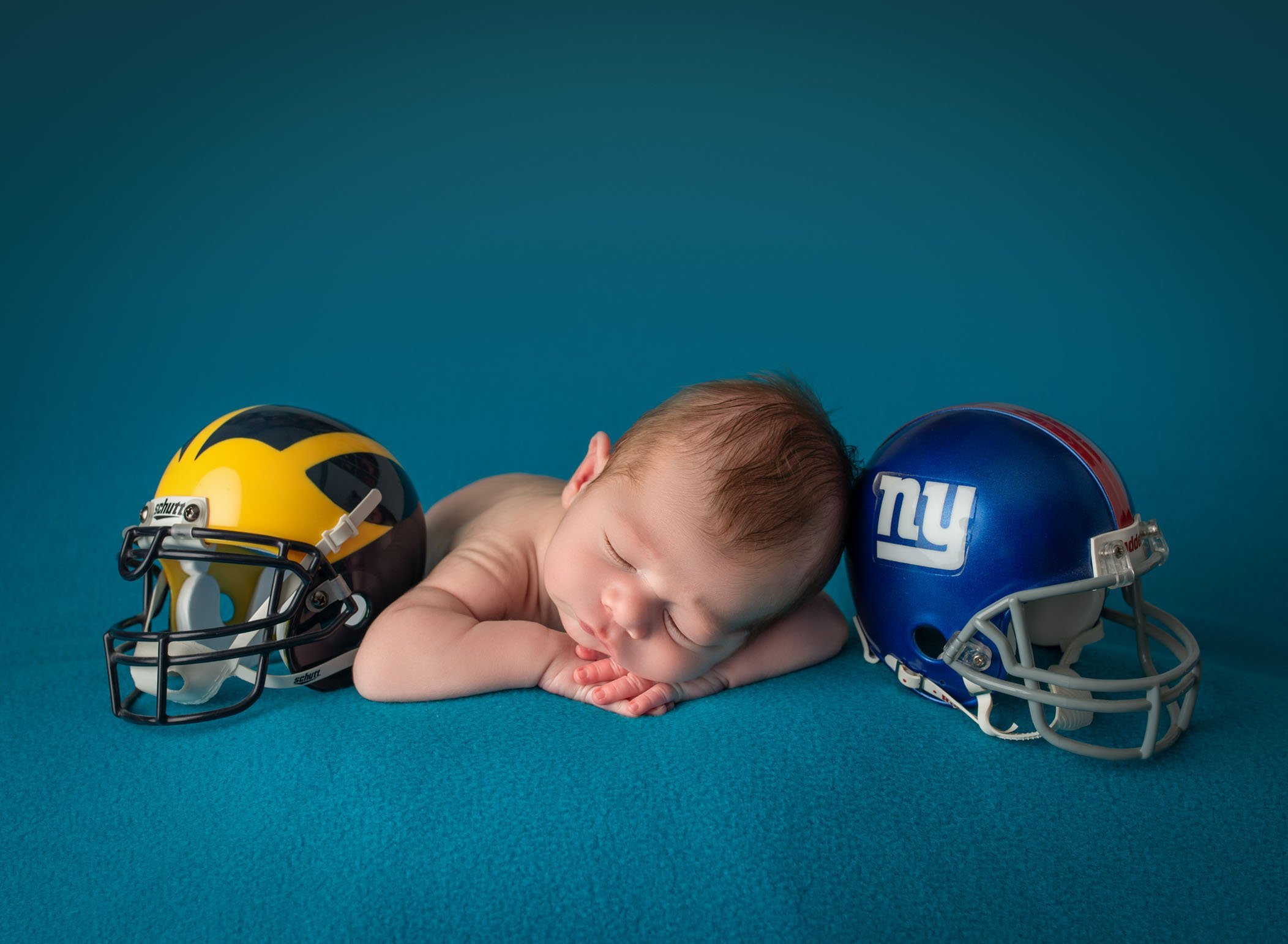 newborn baby boy sleeping on his hands with two football helmets on either side of him