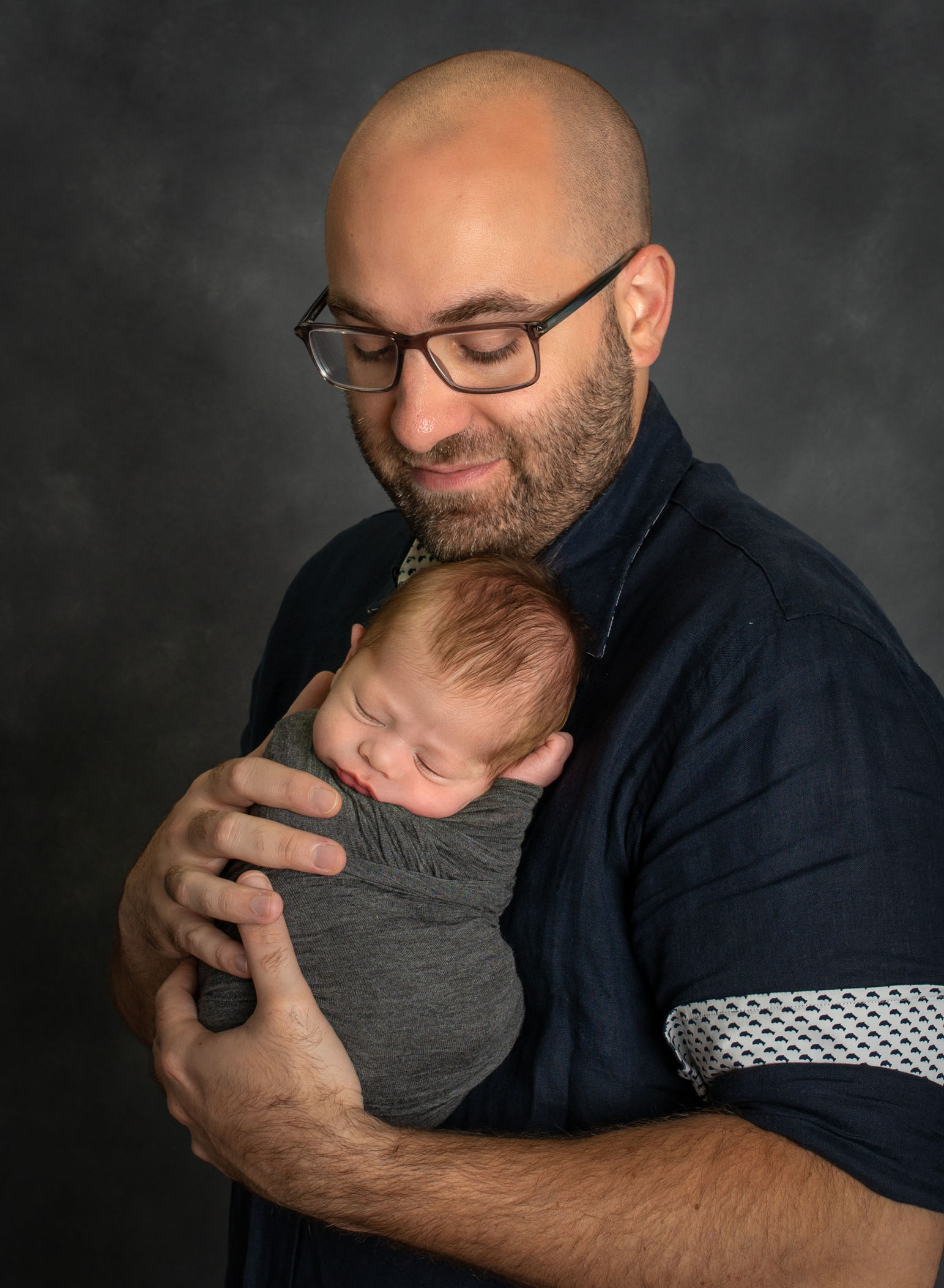 Dad wearing glasses holds his newborn swaddled son against his chest
