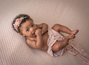 4 month old dark skinned baby girl with her hands in her mouth lying on a pink blanket looking at the camera